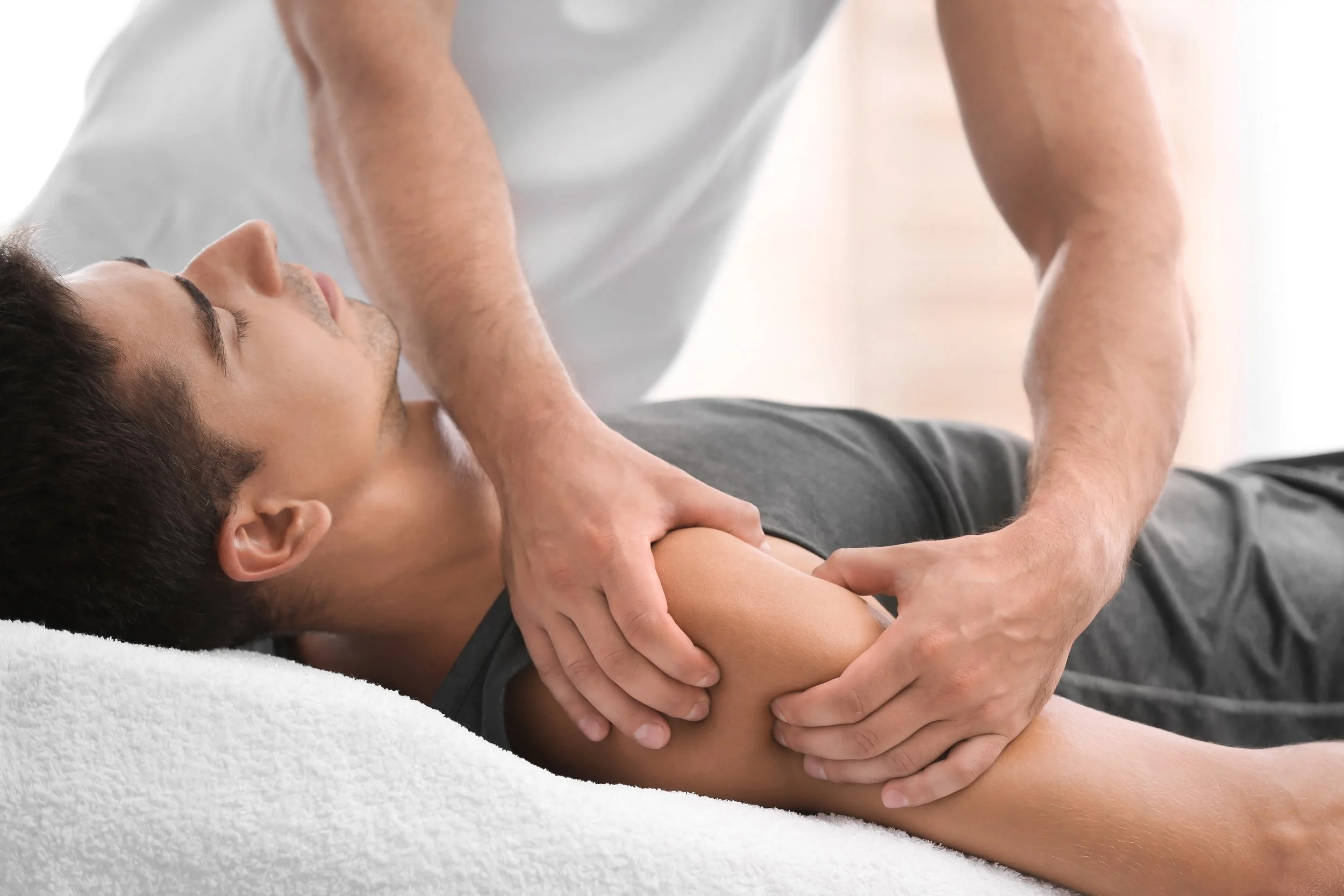 A man receiving a physical therapy or massage on his upper arm from a therapist in a bright, neutral setting.