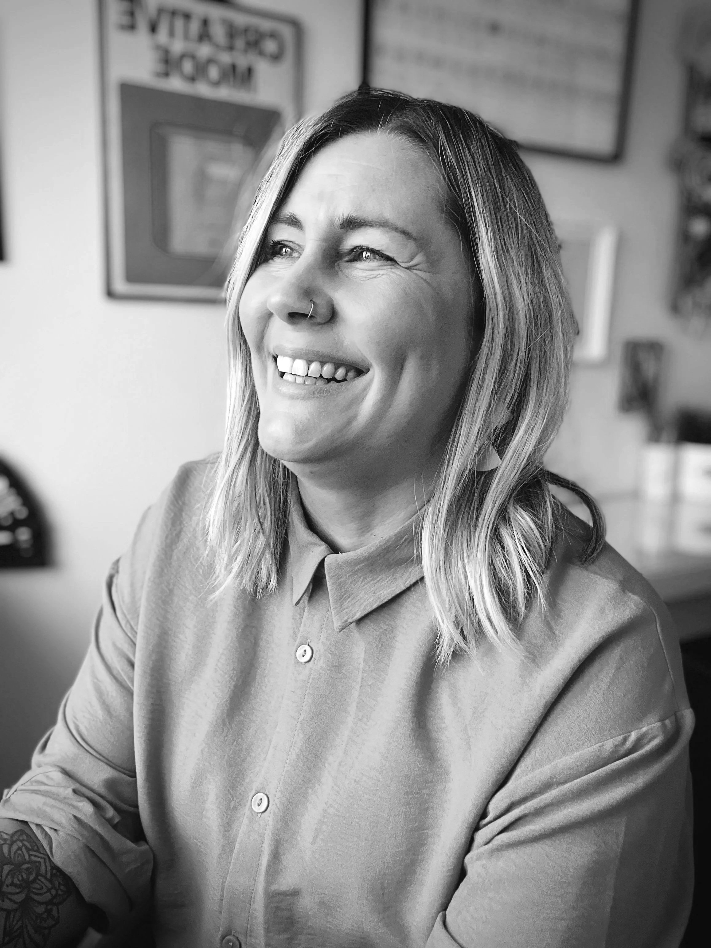 A woman with shoulder-length hair, smiling and looking to her left, wearing a button-up shirt in an indoor setting with framed pictures on the wall behind her.