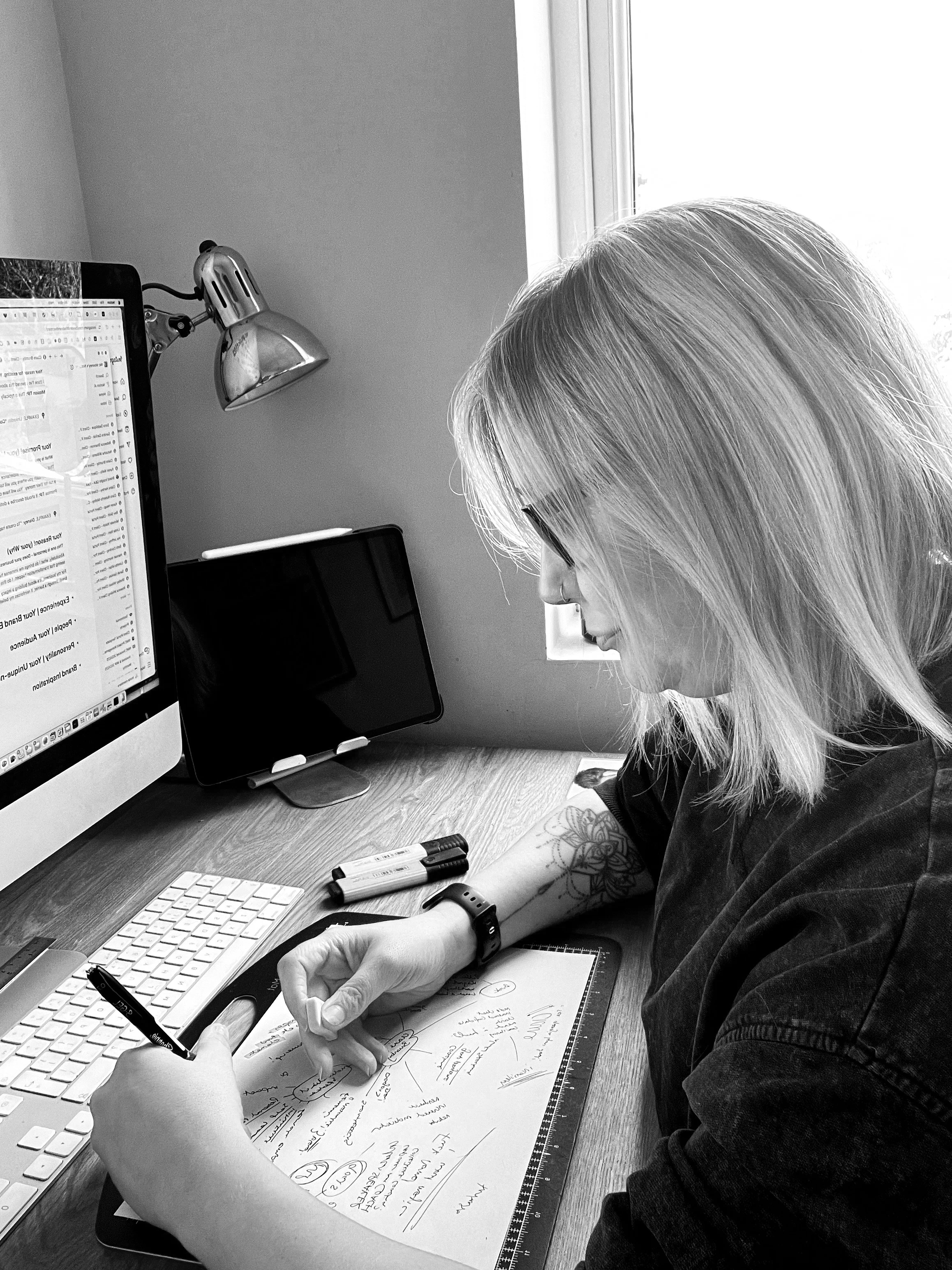 A woman with glasses and shoulder-length hair is sitting at a desk writing on a paper with notes and diagrams, with a computer monitor, keyboard, and a tablet on the desk, near a window.