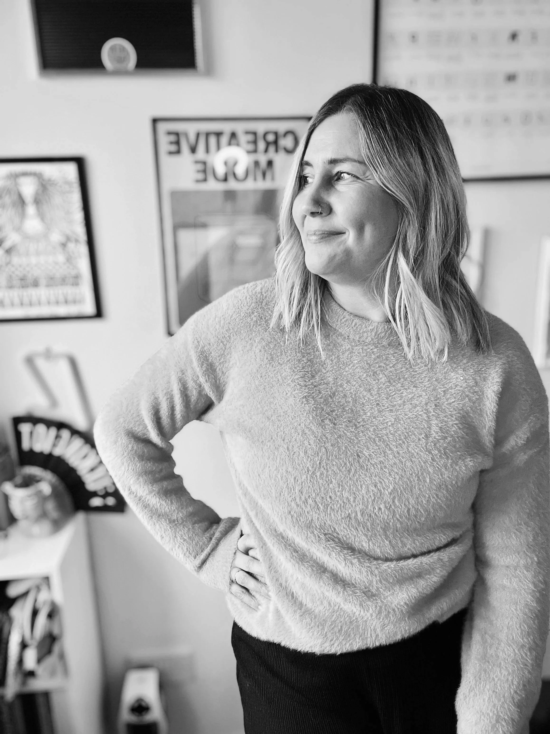A woman with shoulder-length hair, wearing a textured sweater, standing in a room with framed artwork and posters, looking to the side with a slight smile.