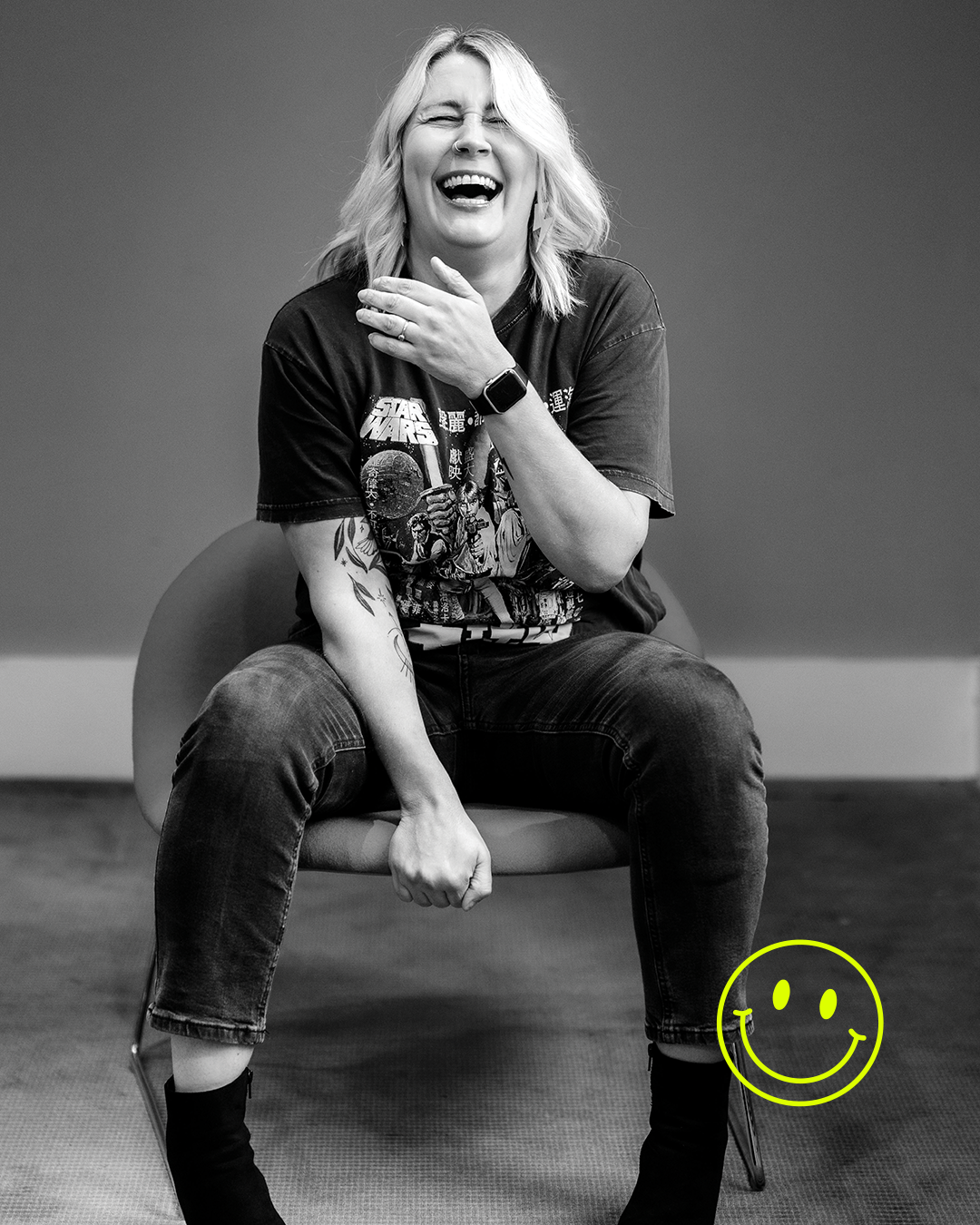 A woman with blond hair, wearing a Star Wars t-shirt and dark jeans, is sitting on a chair, laughing, with her hand on her neck. A yellow smiley face is drawn on the bottom right corner of the image.