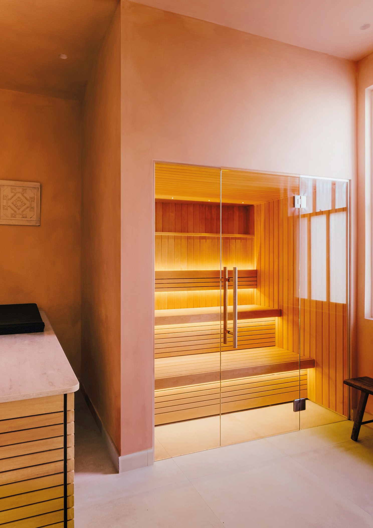 Interior view of a wooden sauna with glass doors, wooden benches, and warm lighting, adjacent to a room with a pink wall and a small marble-top piece of furniture.