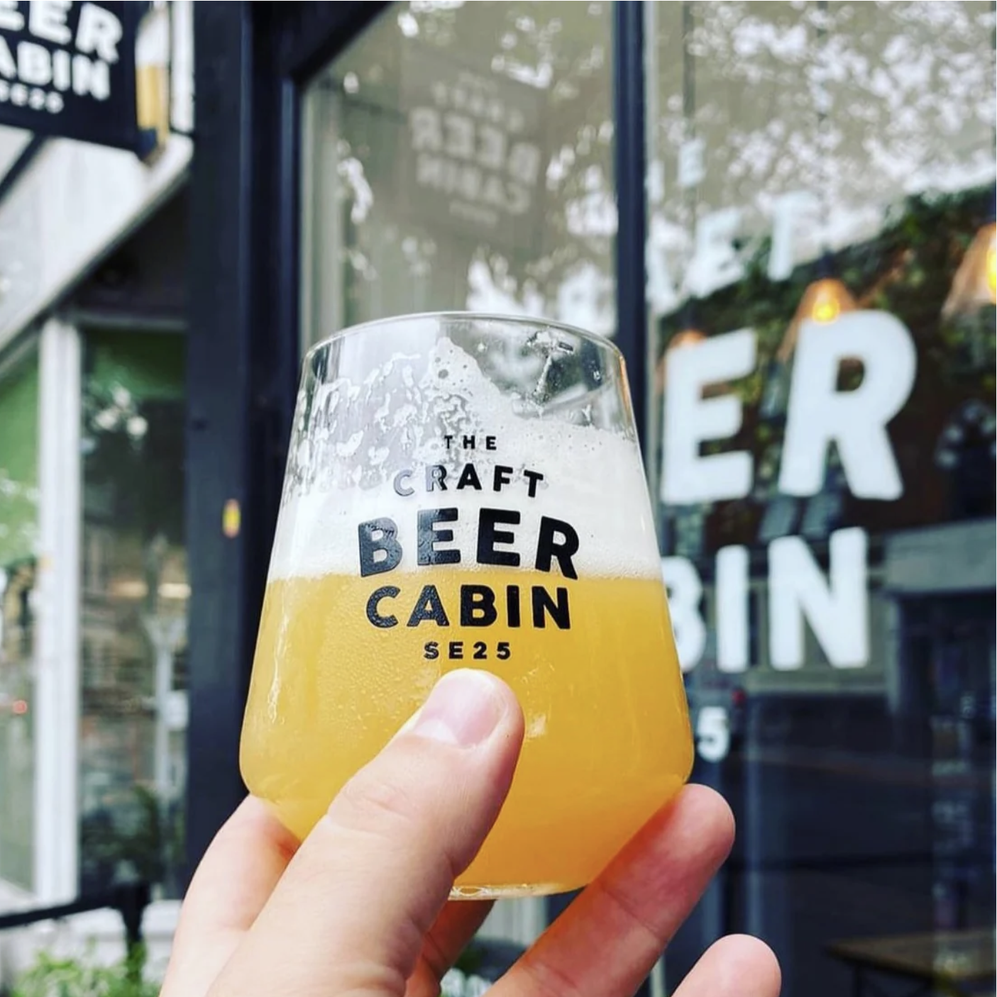 A hand holding a craft beer glass with a foamy head in front of a brewery window with the words 'The Craft Beer Cabin' in the background.
