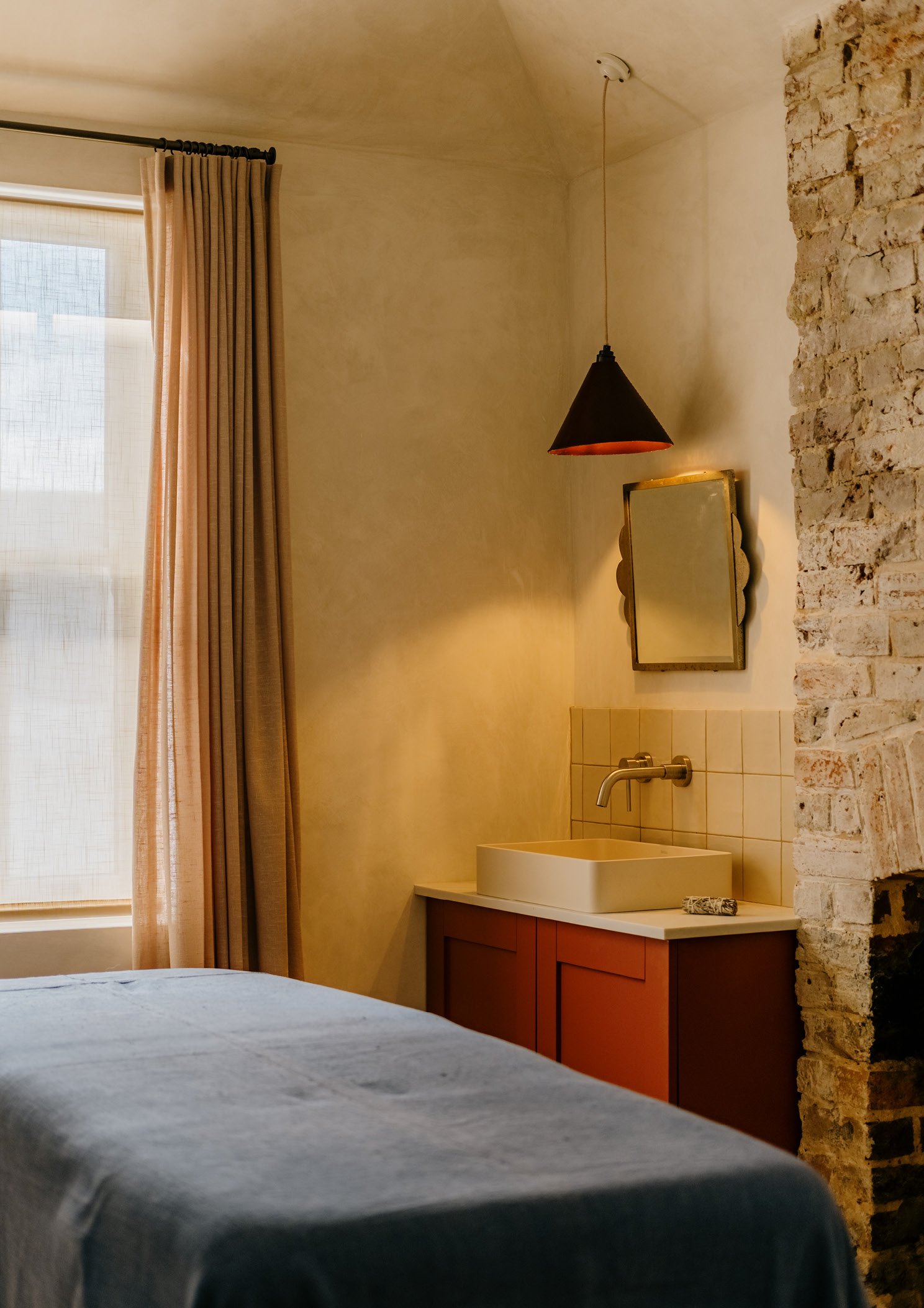 Interior of a bedroom with beige walls, a window with beige curtains, a bed with a dark blue cover, a small sink with a faucet, a mirror above the sink, hanging light fixture, and an exposed brick wall.