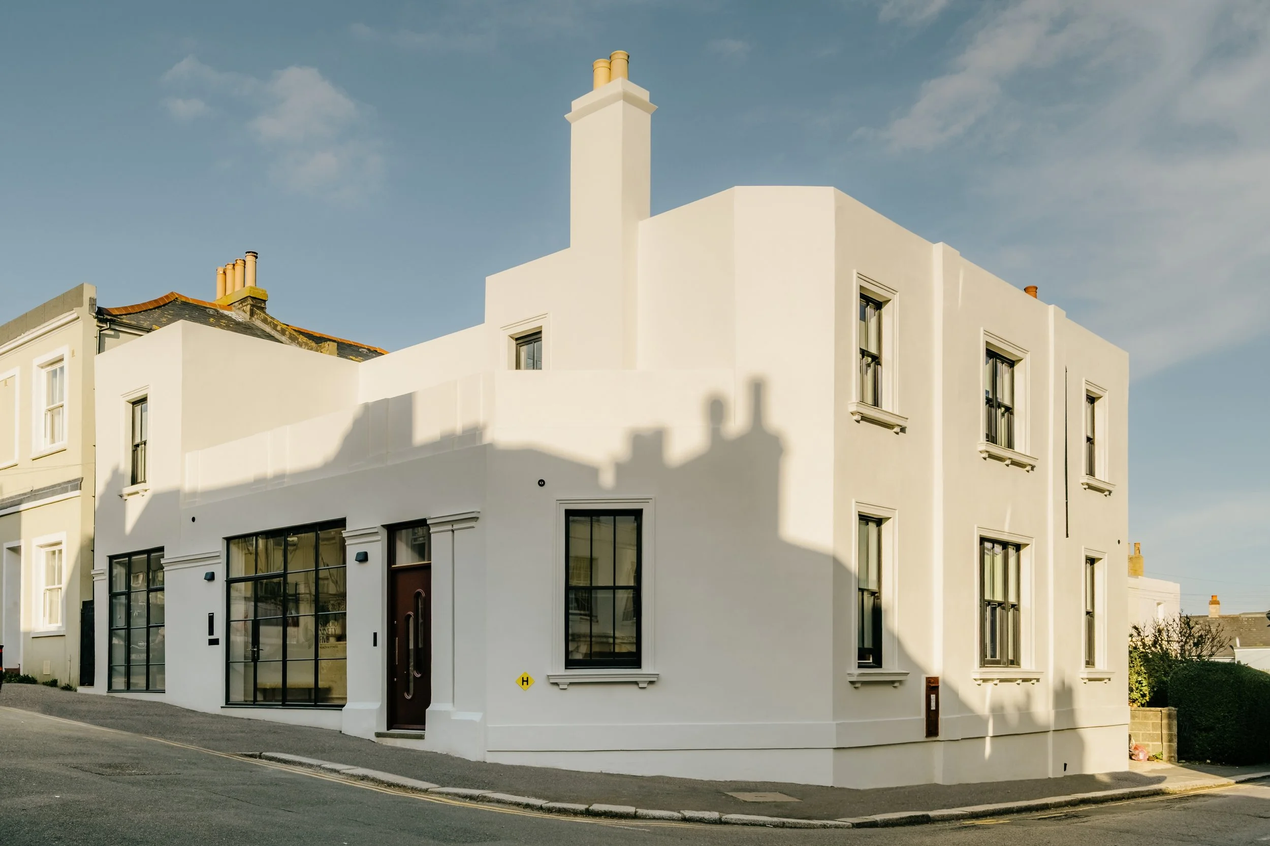 White modern residential building with large windows, situated on a corner street under a blue sky with some clouds.