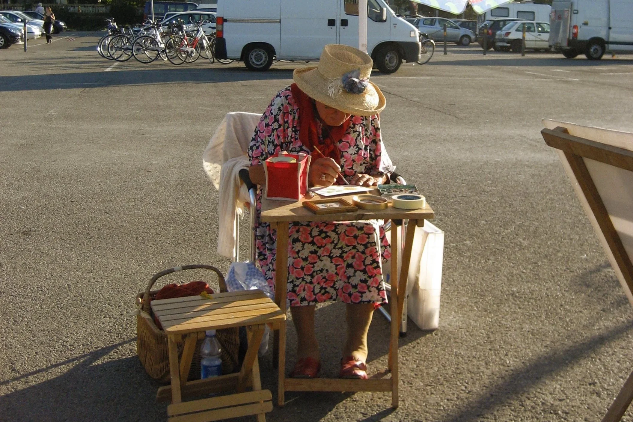 An elderly woman wearing a floral dress, straw hat, and glasses, sitting at a small wooden table on a parking lot. She is writing or drawing on a notepad with various items on the table, including a red container, tape, and framed pictures. There are parked cars, bicycle racks, and vans in the background.