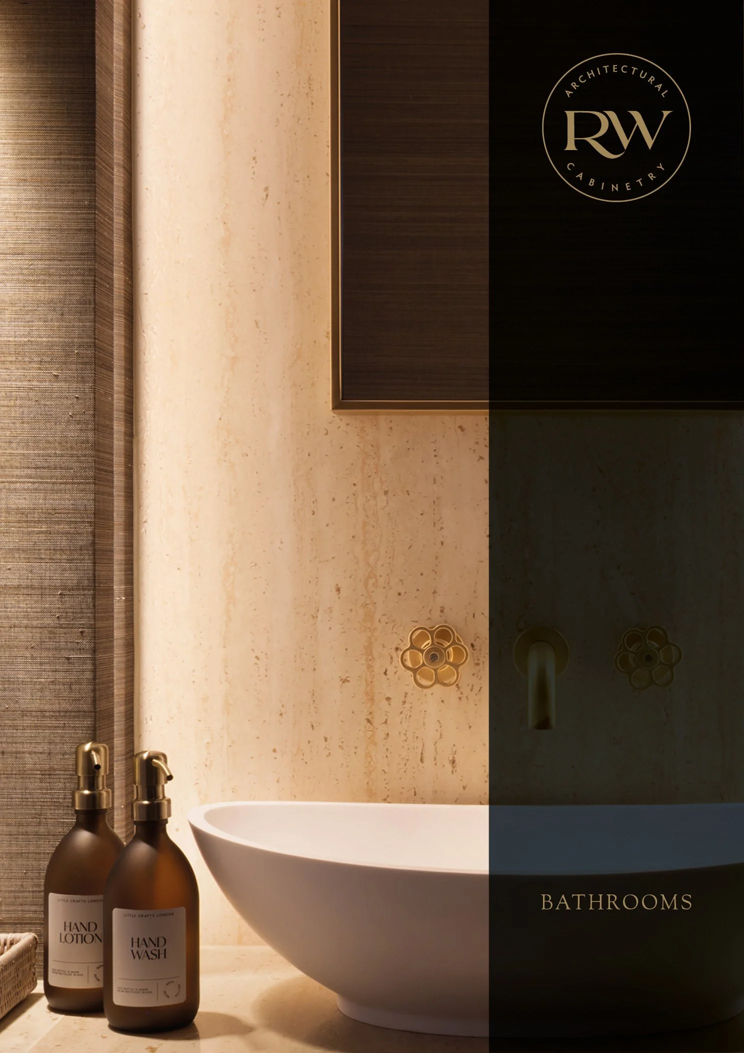 A modern bathroom with a beige stone wall, a white vessel sink, gold fixtures, and two brown bottles labeled hand lotion and hand wash on a beige countertop.
