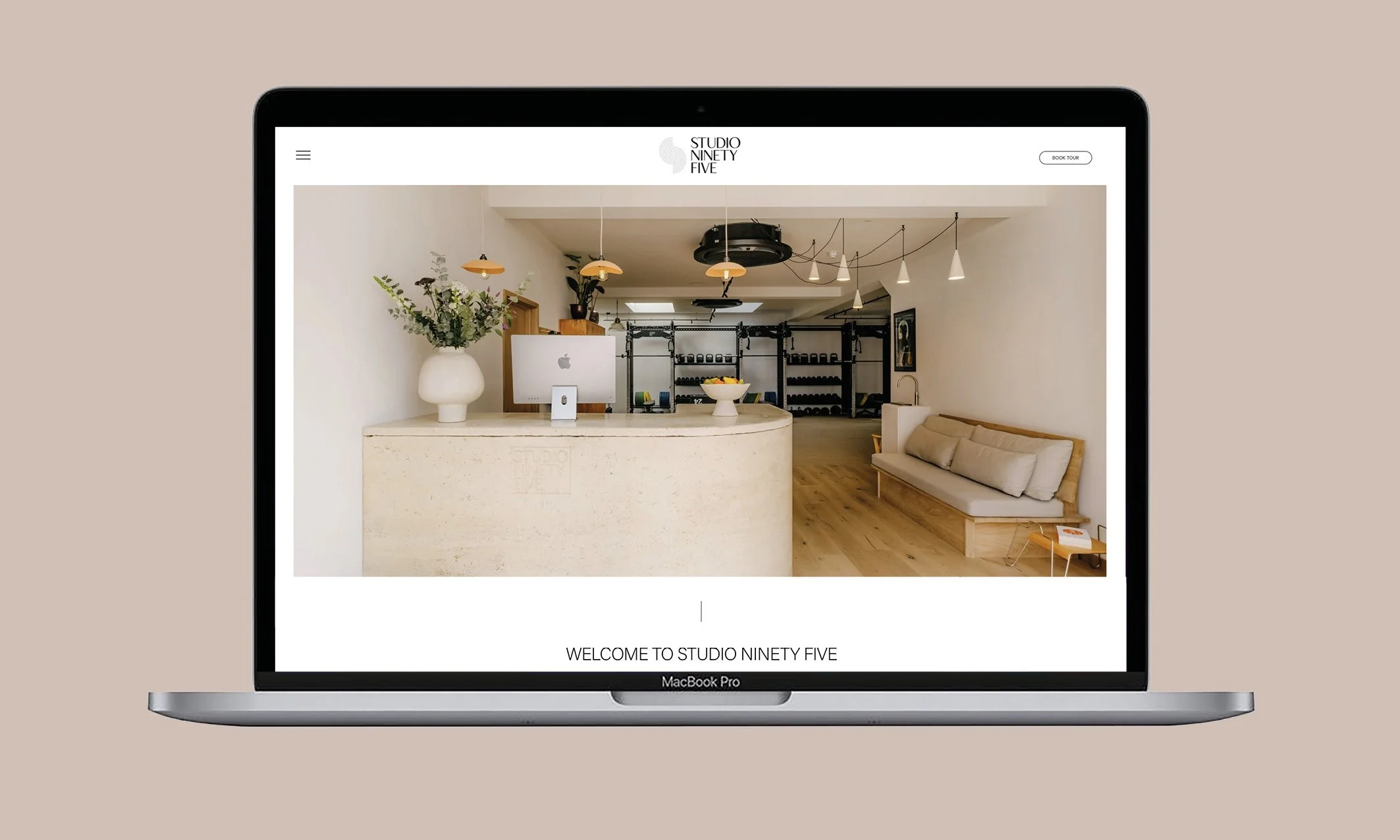 Laptop displaying the homepage of Studio Ninety Five, featuring a reception area with a beige countertop, potted plant, computer, bowl of fruit, a beige sofa, and exercise equipment in the background.