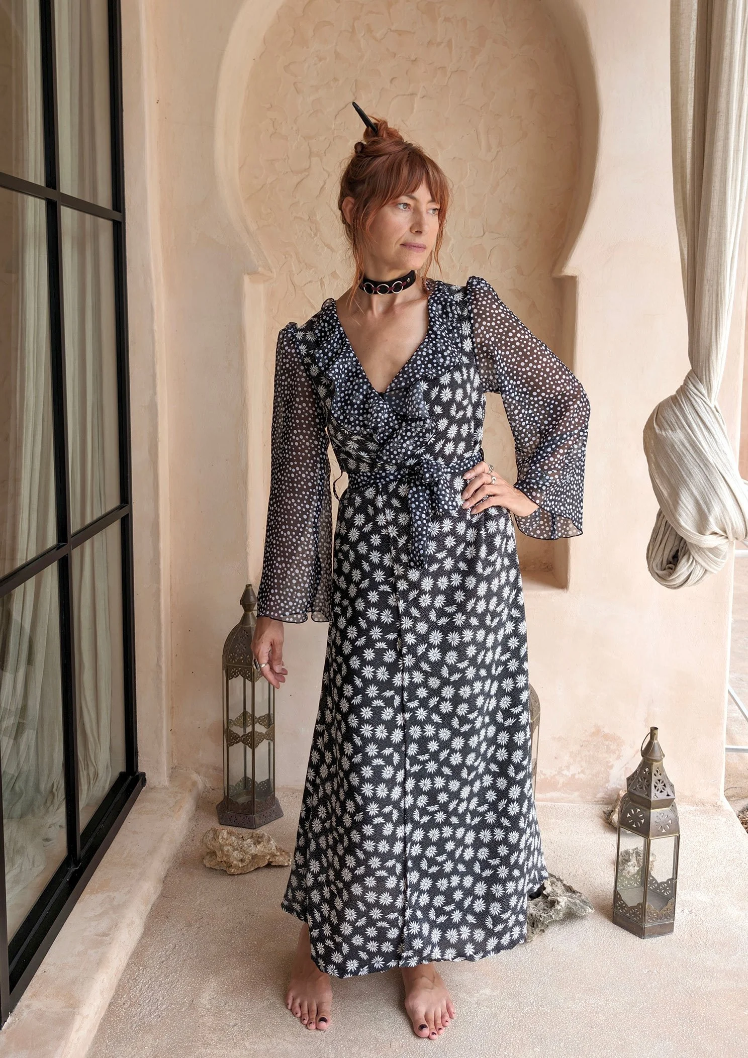 A woman with red hair wearing a black and white floral dress, standing barefoot indoors near a window with stone lanterns on the floor.
