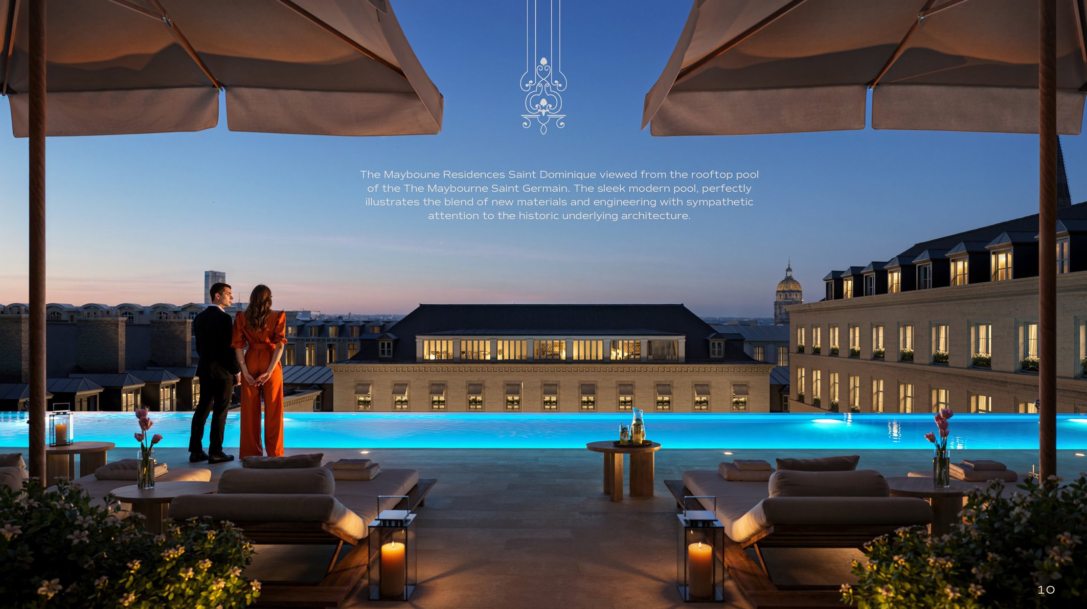 A rooftop pool at dusk with a couple standing and talking, overlooking city buildings and a dome. The pool has modern lighting and the area is decorated with flowers, candles, and lounge chairs.