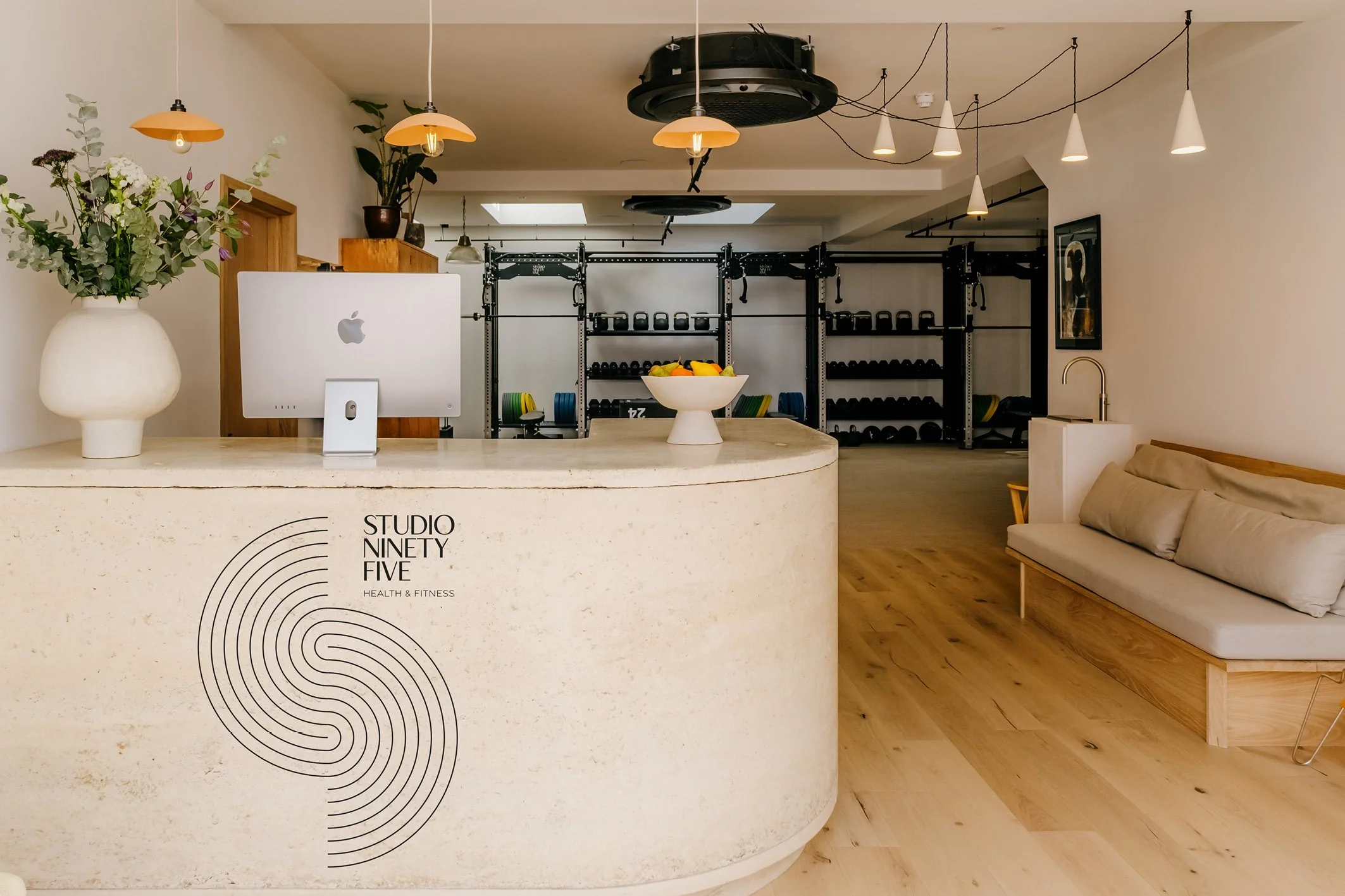 Reception area of a health and fitness studio named Studio Ninety Five with a curved beige desk, a computer, a bowl of fruit, and a bench with cushions. In the background, weightlifting equipment and kettlebells are visible, with modern pendant lighting and wooden accents.