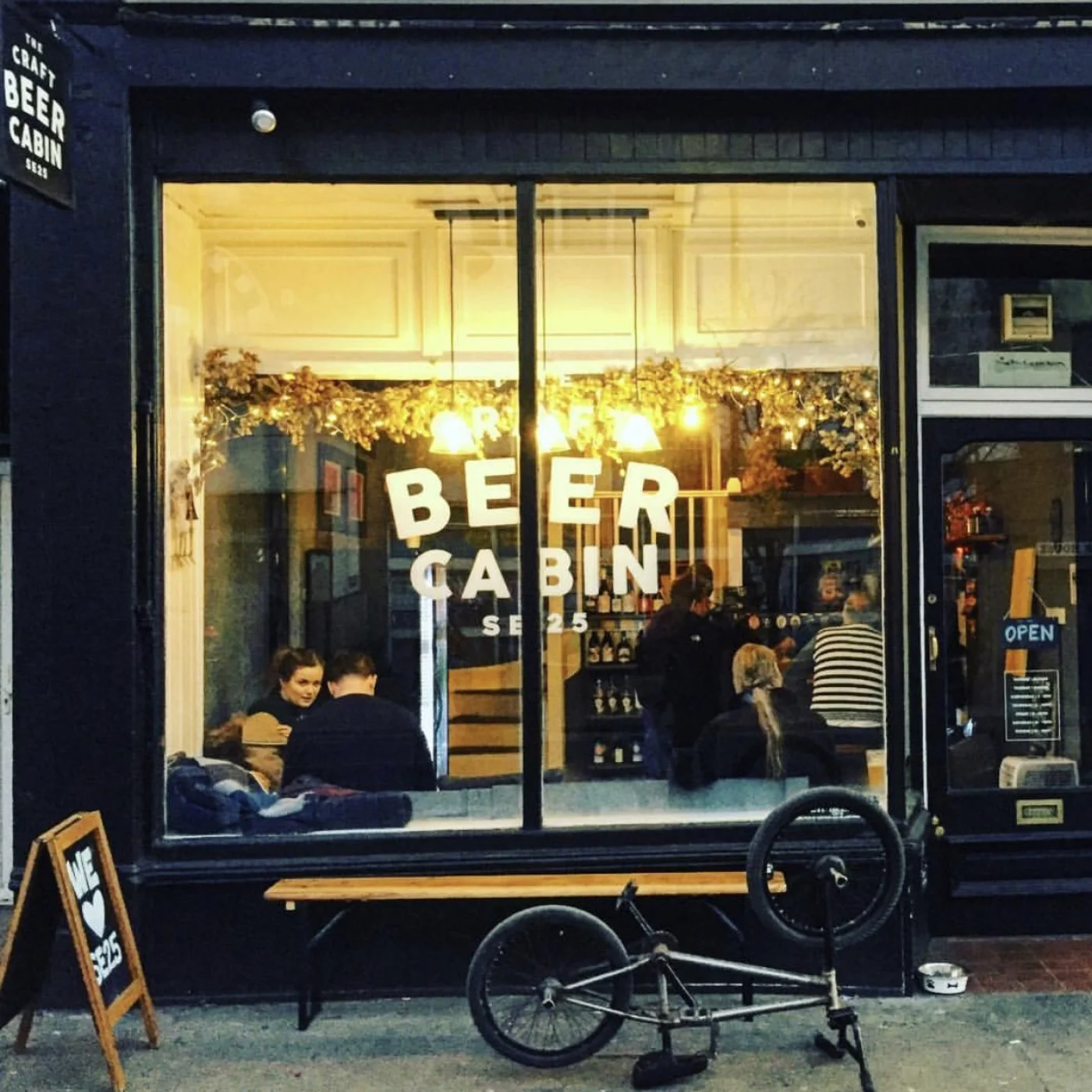 View of a craft beer bar called 'Beer Cabin' with large front window showing customers inside, hanging lights, and sign outside advertising $2.50 beer.