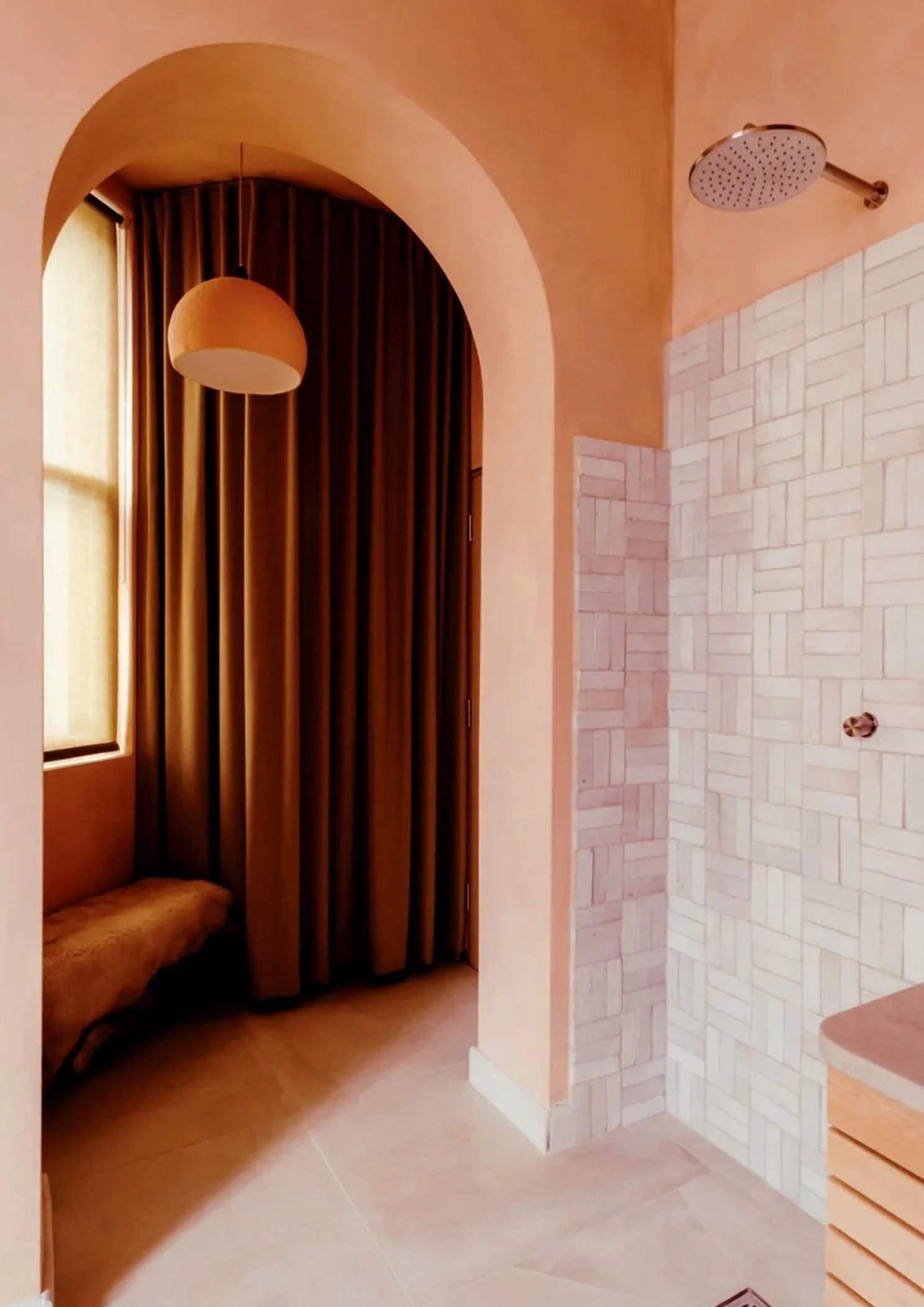 A bathroom with arched doorway, featuring a showerhead on patterned wall, a hanging pendant light, a window with a yellow curtain, and a bench near the window.