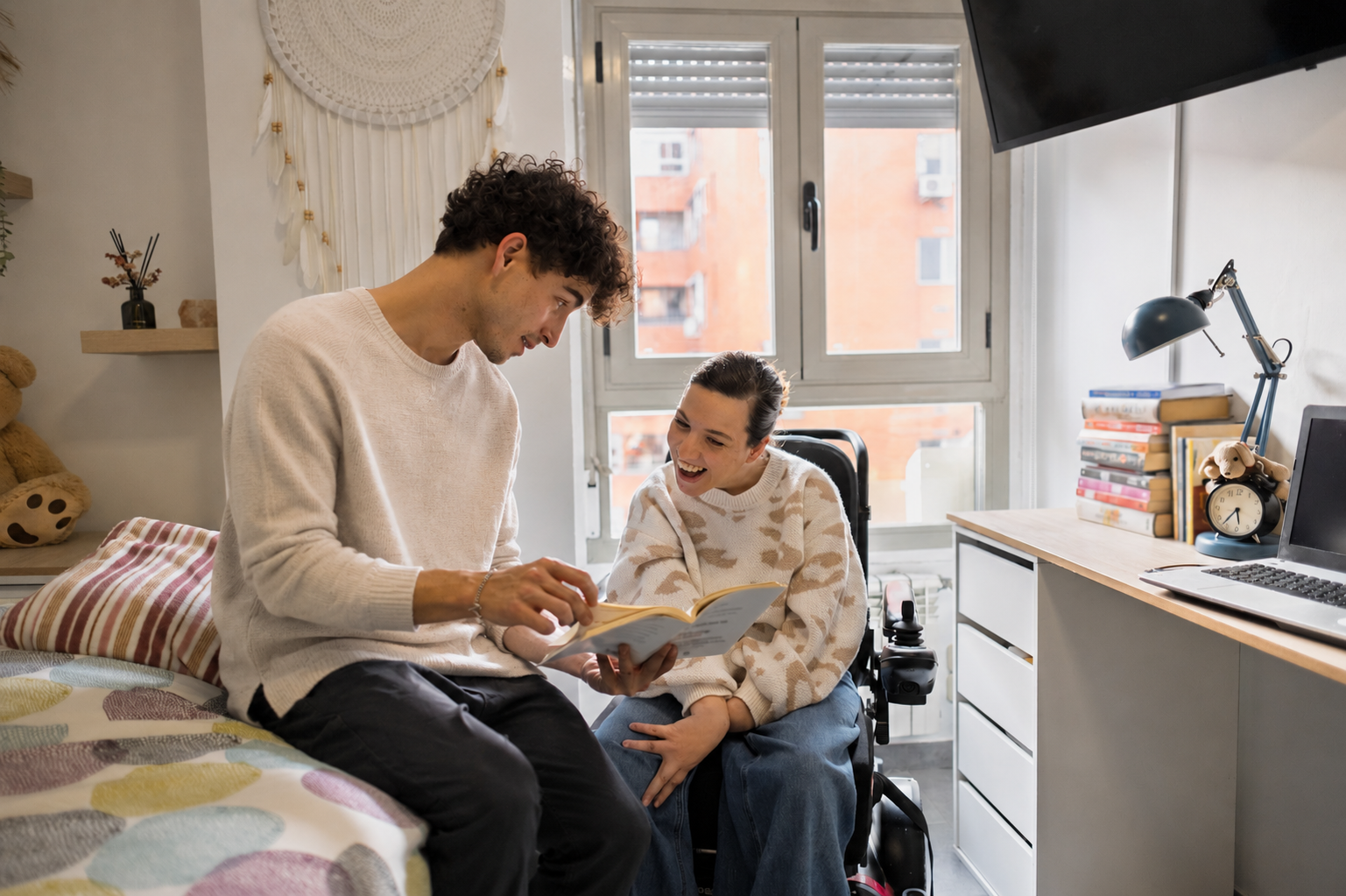 A young man and a young woman in a bedroom, sitting on a bed with colorful blanket, looking at a book and smiling.