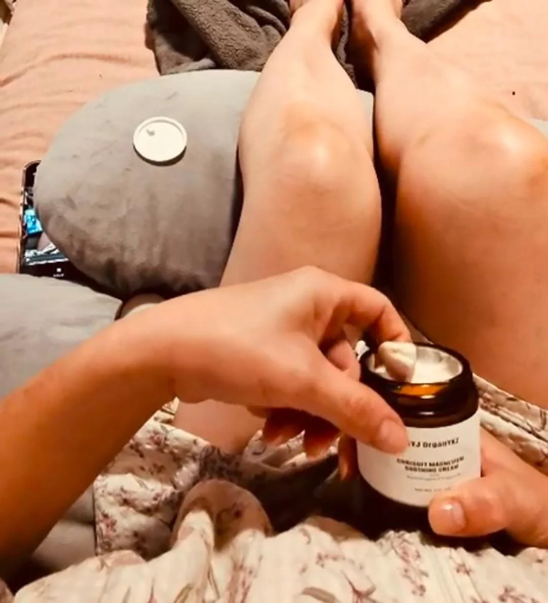 Person sitting on a couch with legs extended, holding an open supplement bottle with a spoon inside, and a candle placed nearby on a cushion.