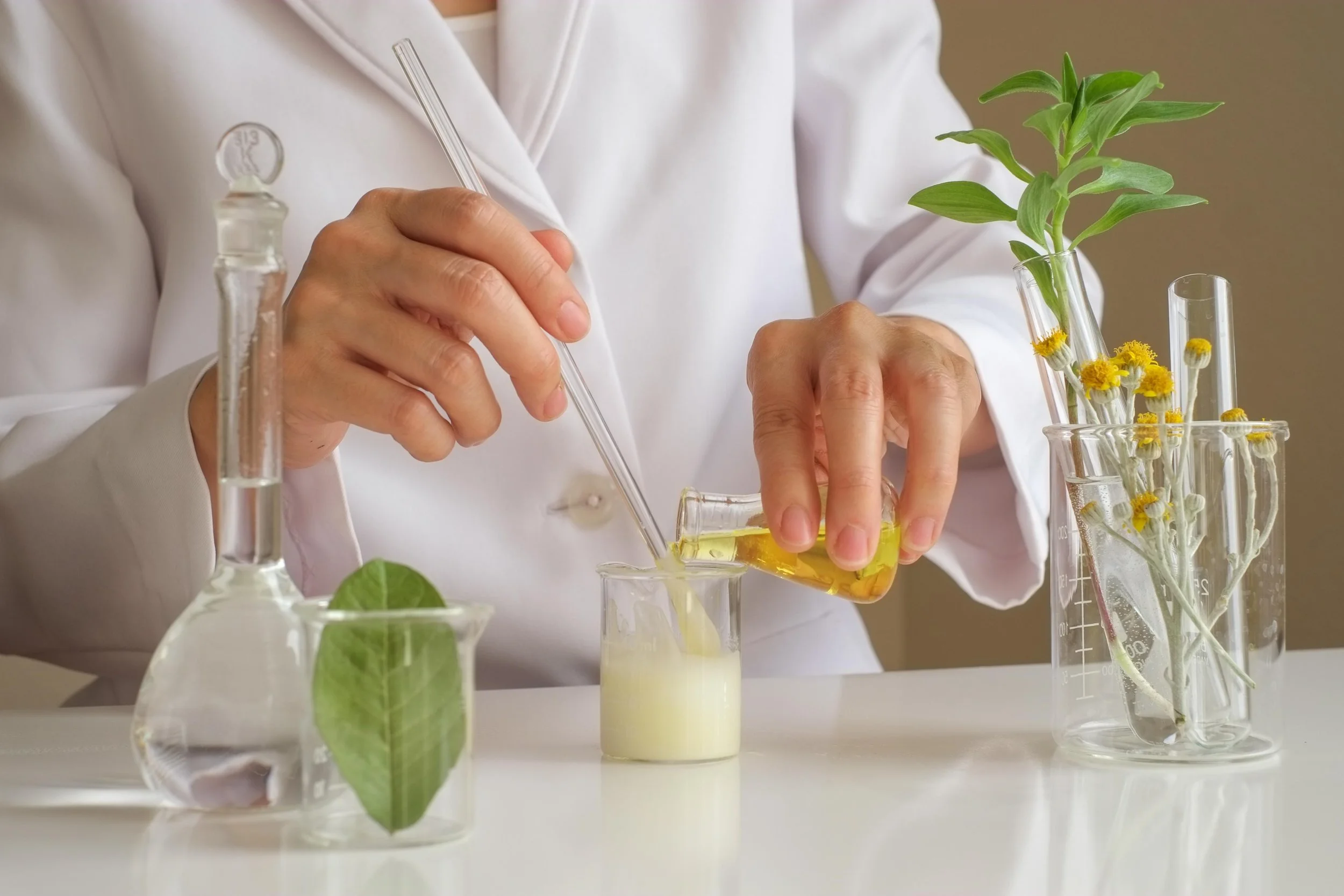 Close-up of a scientist in a white lab coat conducting experiments with test tubes containing yellow liquid, green plants, and small flowers in a laboratory setting.