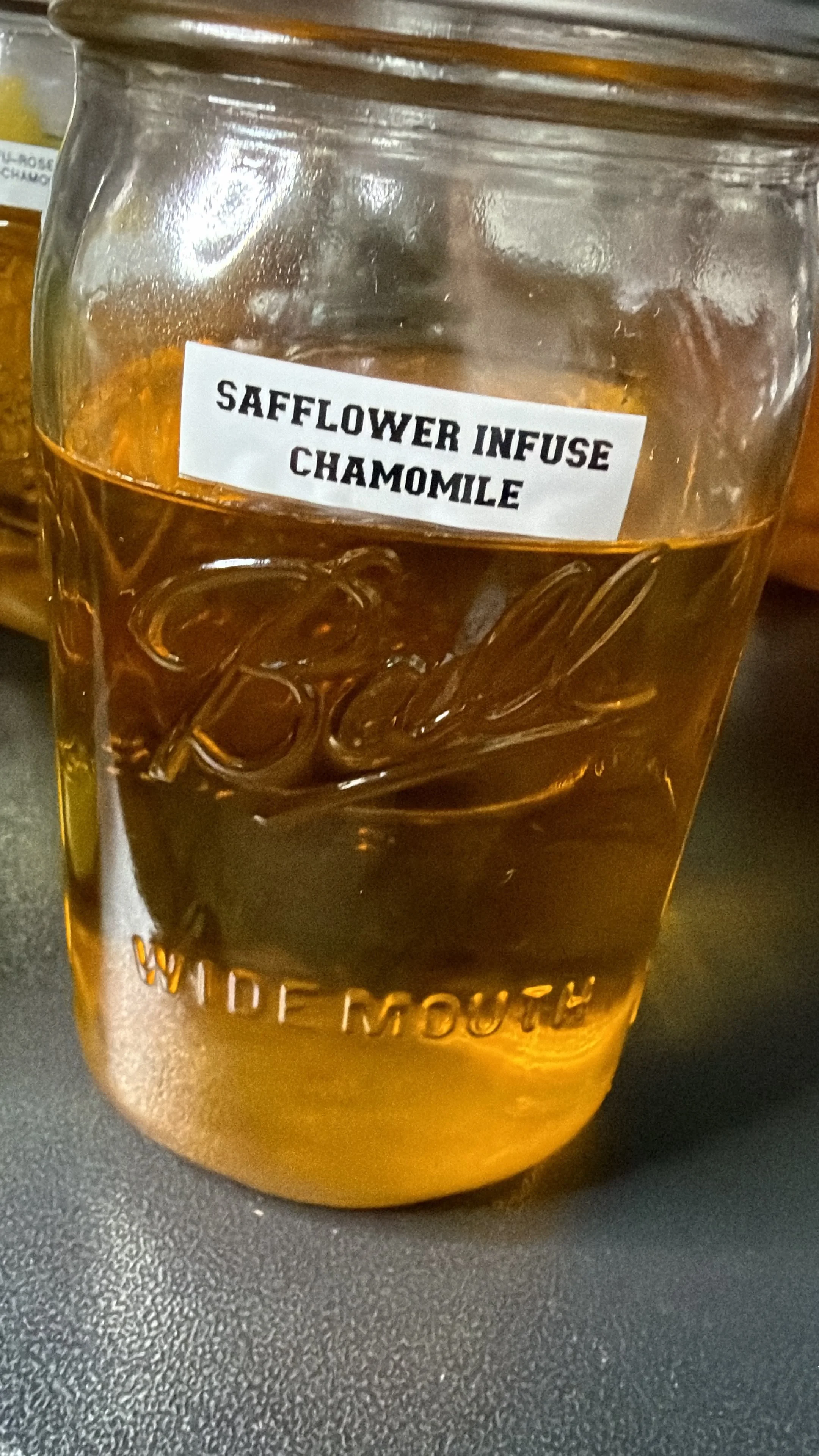 A glass jar labeled 'Safflower Infuse Chamomile' filled with a yellow-brown liquid, sitting on a black textured surface.
