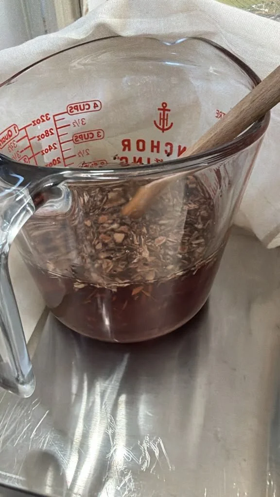 A glass measuring cup with red markings containing a dark liquid and wood chips, with a wooden spoon inside, placed on a metallic surface.