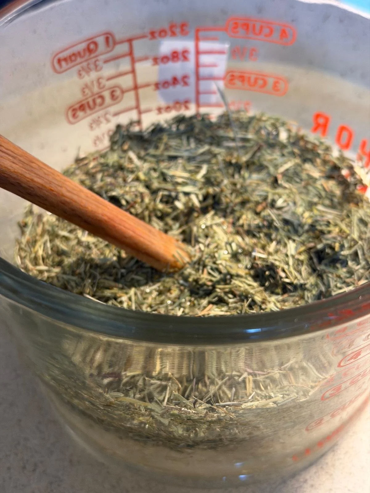Close-up of a clear measuring cup filled with dried, chopped herbs and a wooden spoon inside.