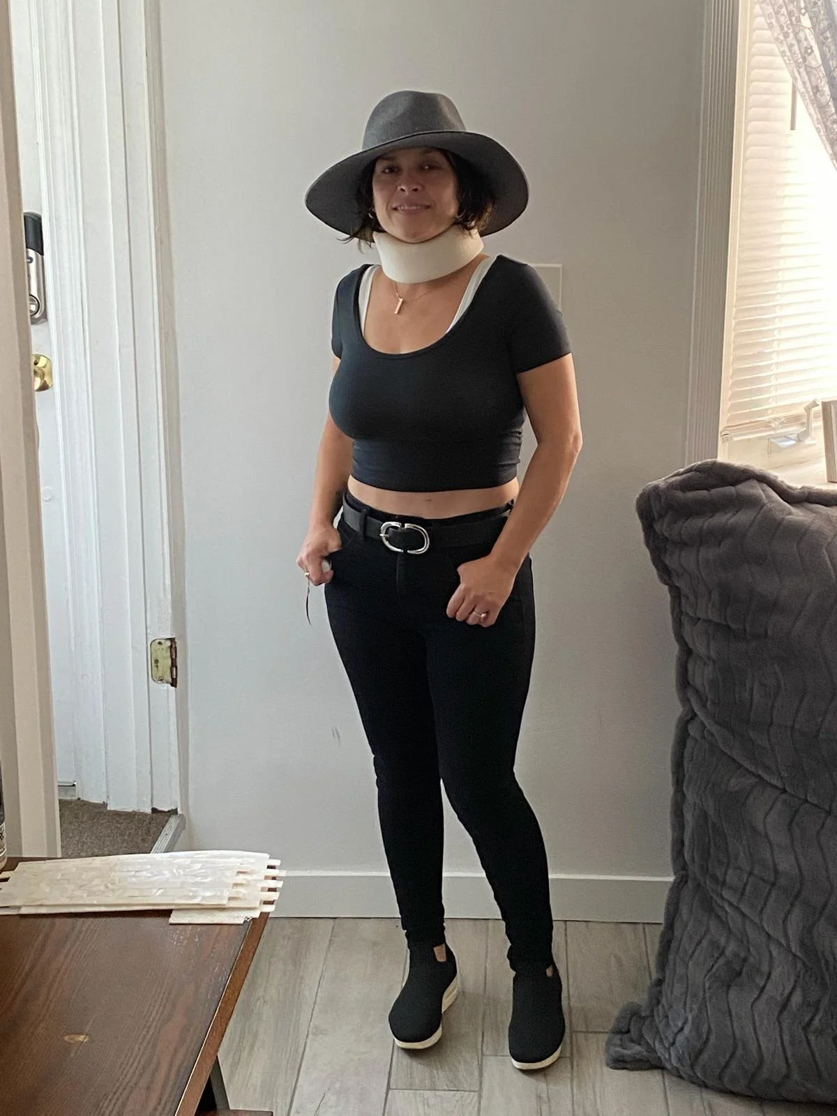 Woman standing indoors in front of a white wall, wearing a wide-brimmed gray hat, black crop top, high-waisted jeans with a belt, black sneakers, and a neck brace.