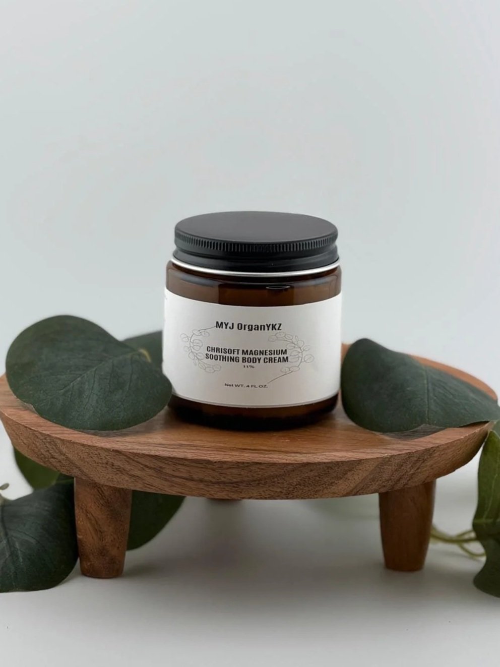 A brown jar with a white label and black lid sits on a small wooden surface, surrounded by green leaves.