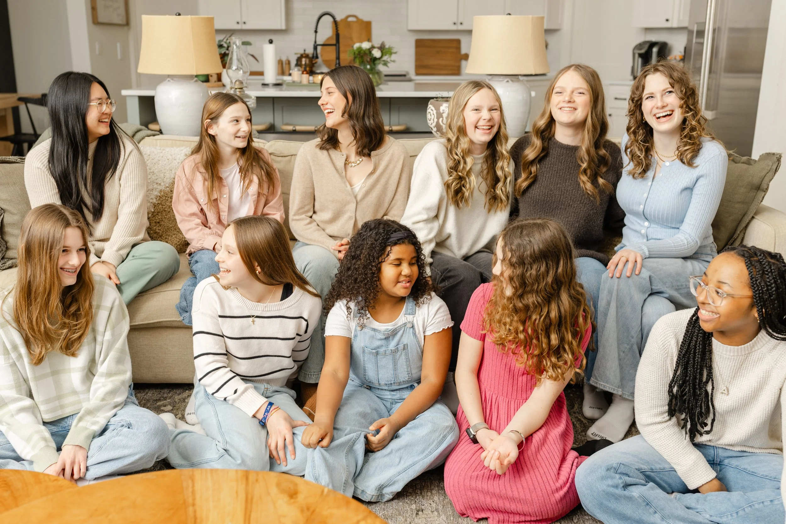 A group of children and women sitting on a carpeted floor in a living room, laughing and enjoying each other's company.