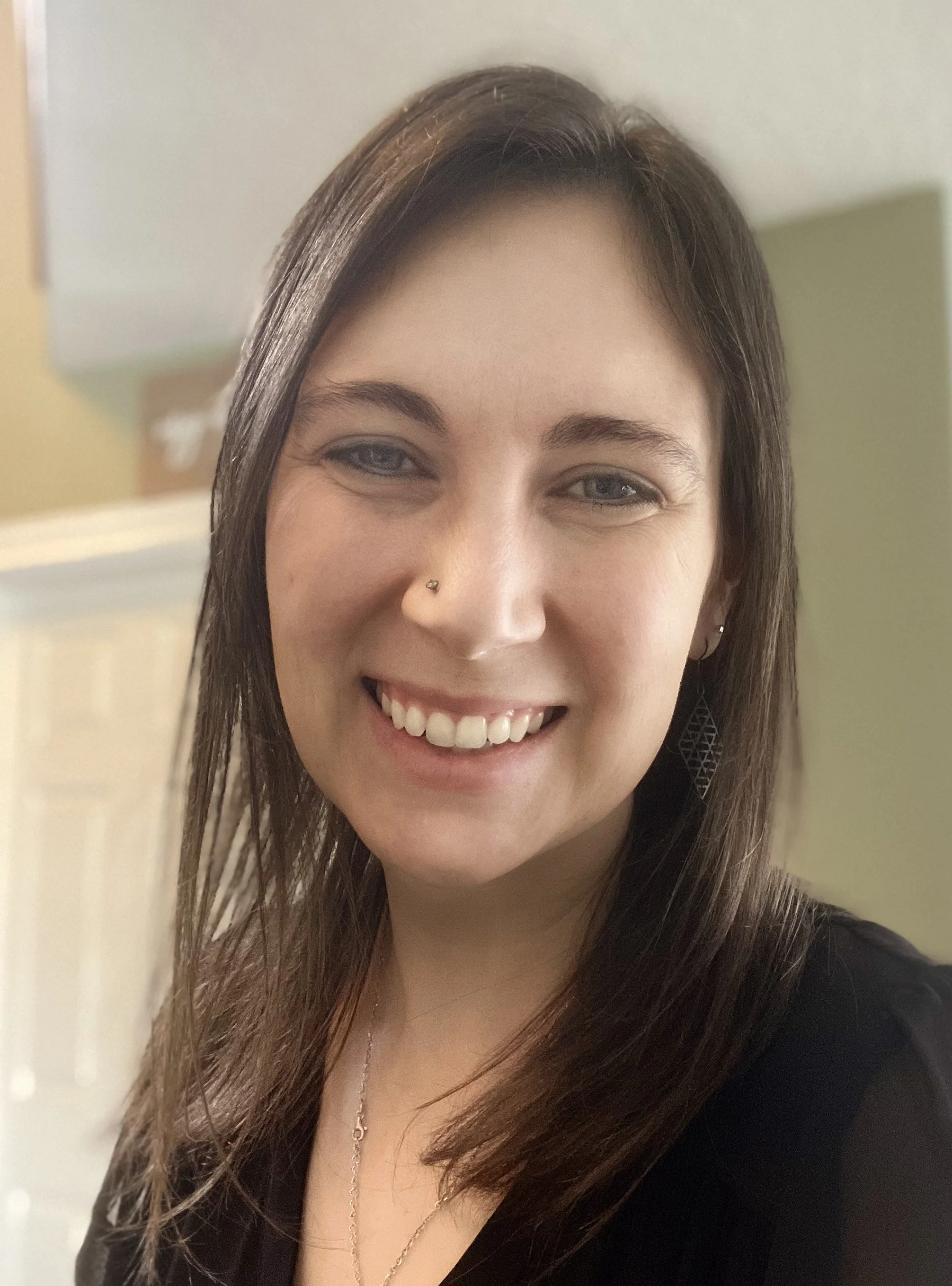 A woman with brown hair and blue eyes smiling at the camera, wearing a black top, earrings, and a nose piercing, indoors with a green wall and door in the background.