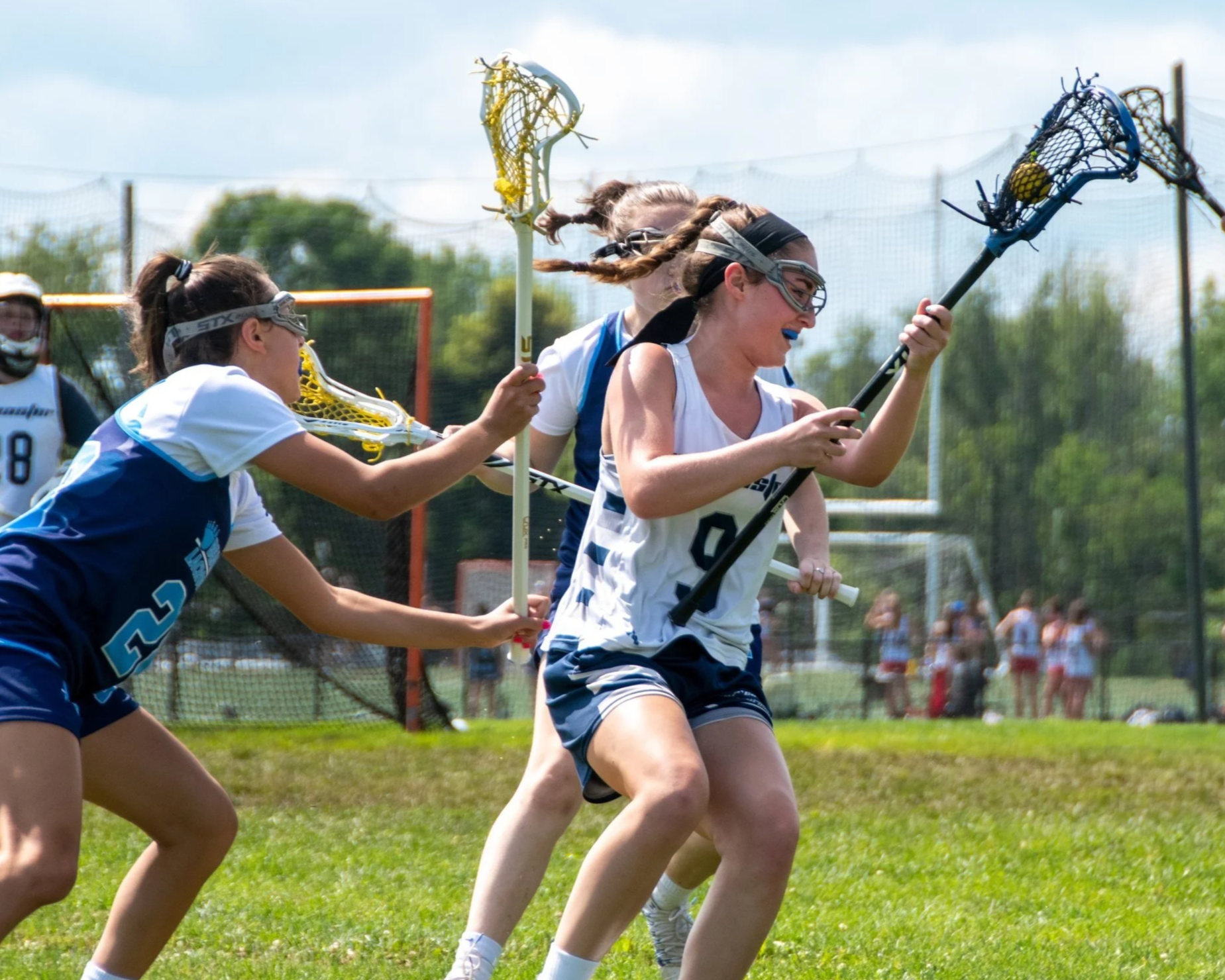 Girls playing lacrosse outdoors on a grassy field during daytime, wearing sports uniforms and goggles.