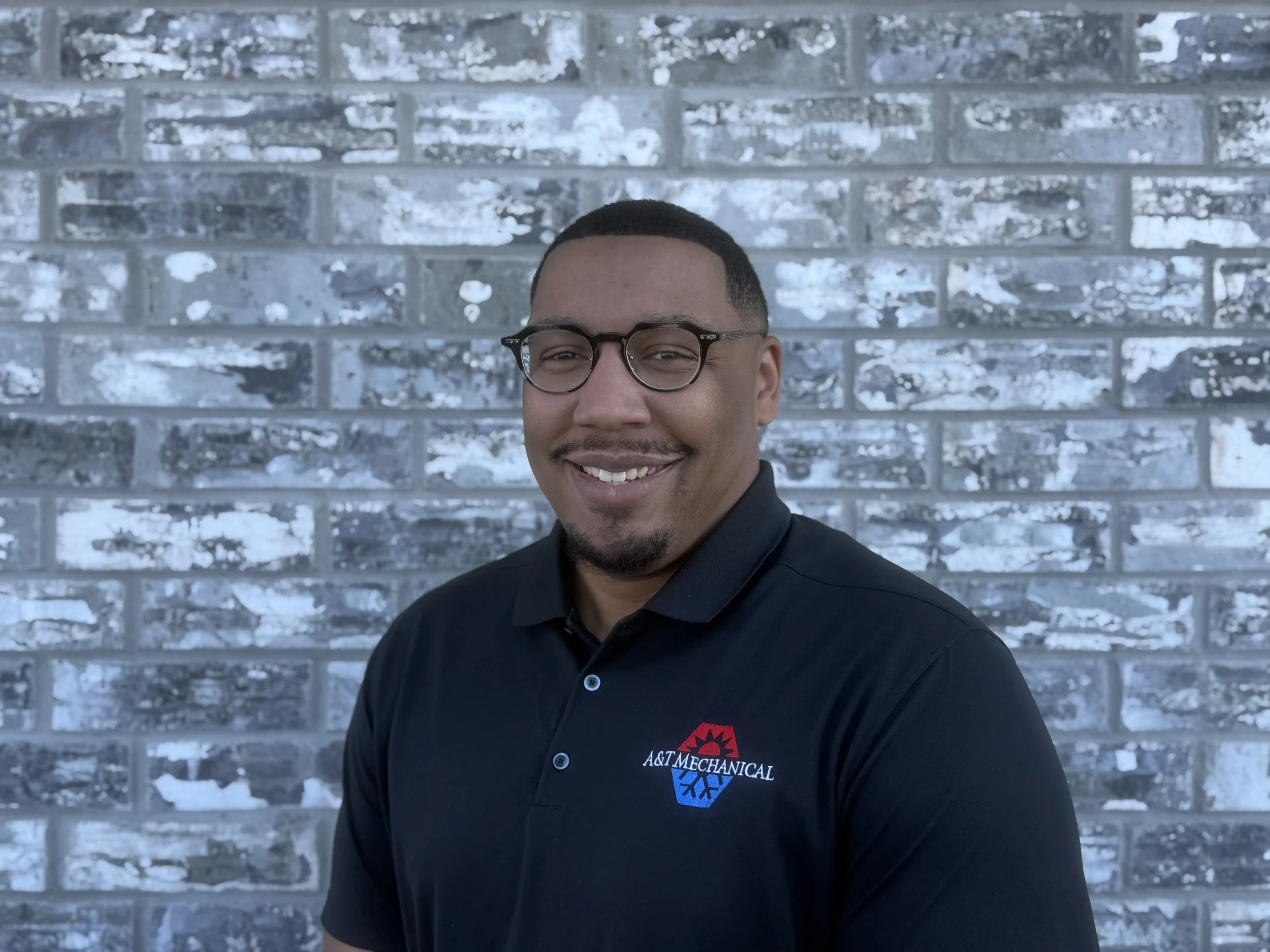 Portrait of a smiling man wearing a red polo shirt with the logo 'A&T Mechanical Heat and Air Services' on the chest, in front of a textured blue-gray background.