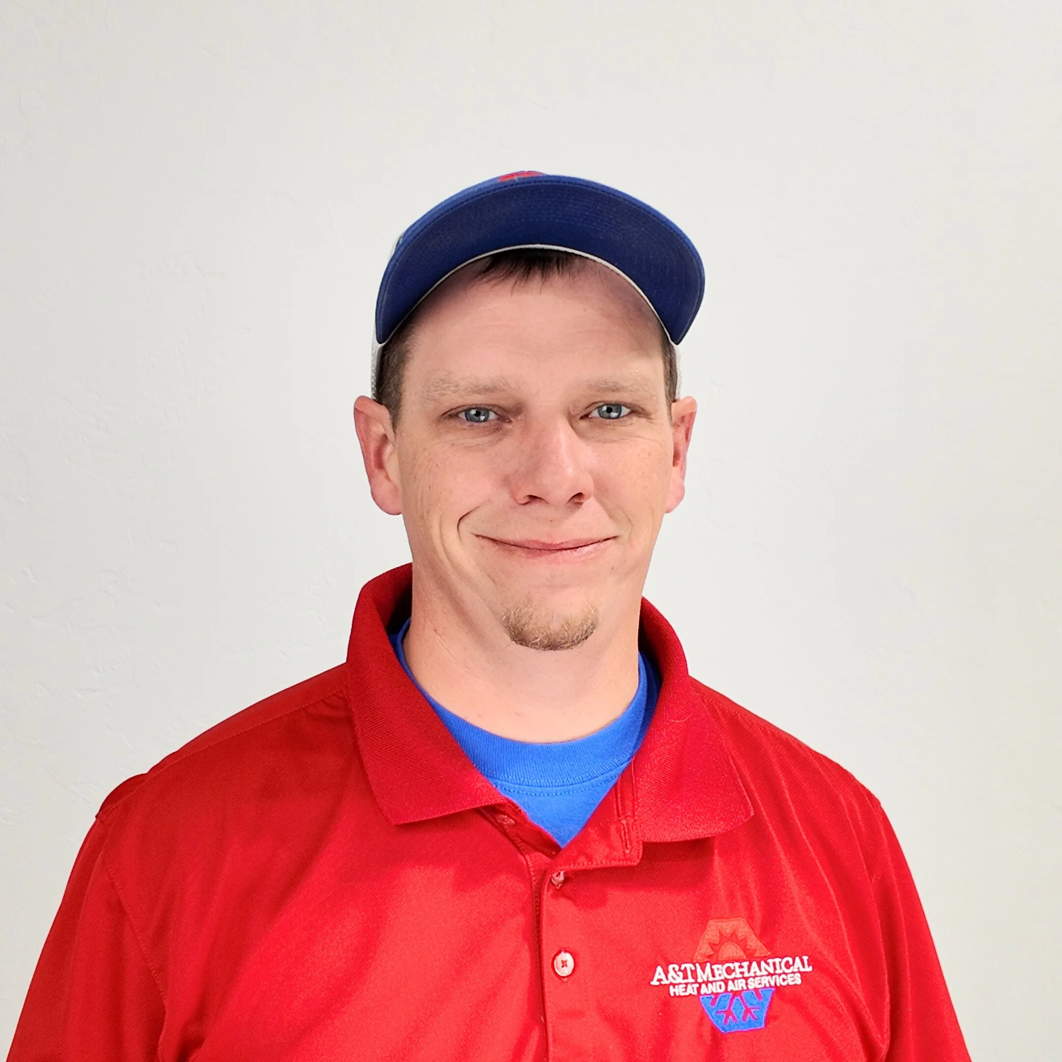 A man wearing a gray and black cap, a red polo shirt with the logo of A&M Mechanical Heat and Air Services, smiling at the camera, against a textured blue background.