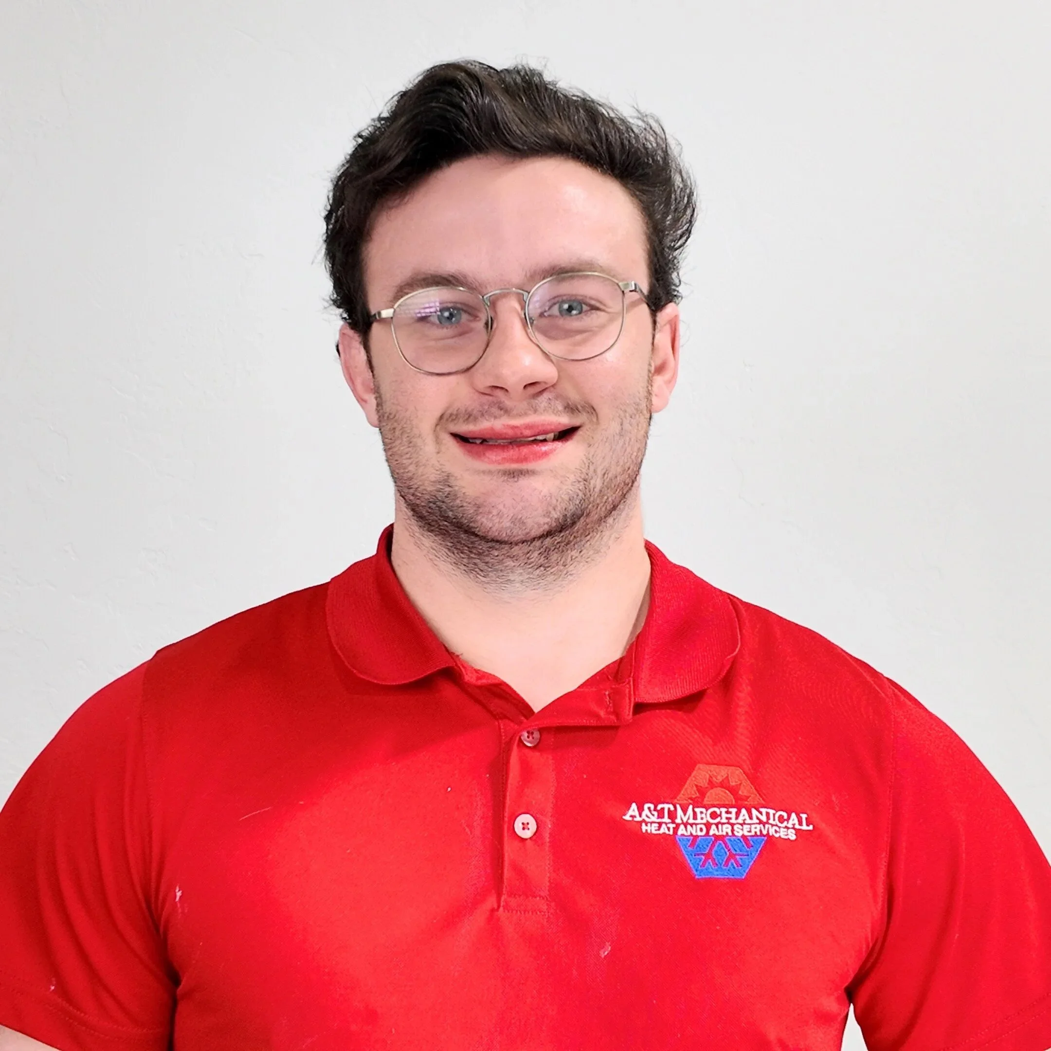 A young man with curly dark hair, glasses, and a mustache, wearing a red polo shirt with the logo for A&T Mechanical, a heating and air conditioning business.