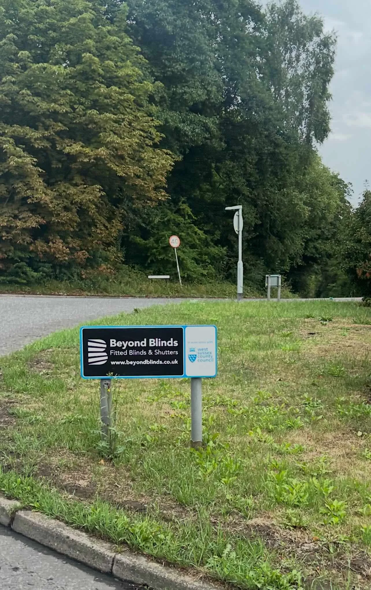 A roadside sign advertising Beyond Blinds, a company that sells fitted blinds and shutters, with the website beyondblinds.co.uk, located near a grassy area and a road with a 40 mph speed limit sign in the background, surrounded by trees.