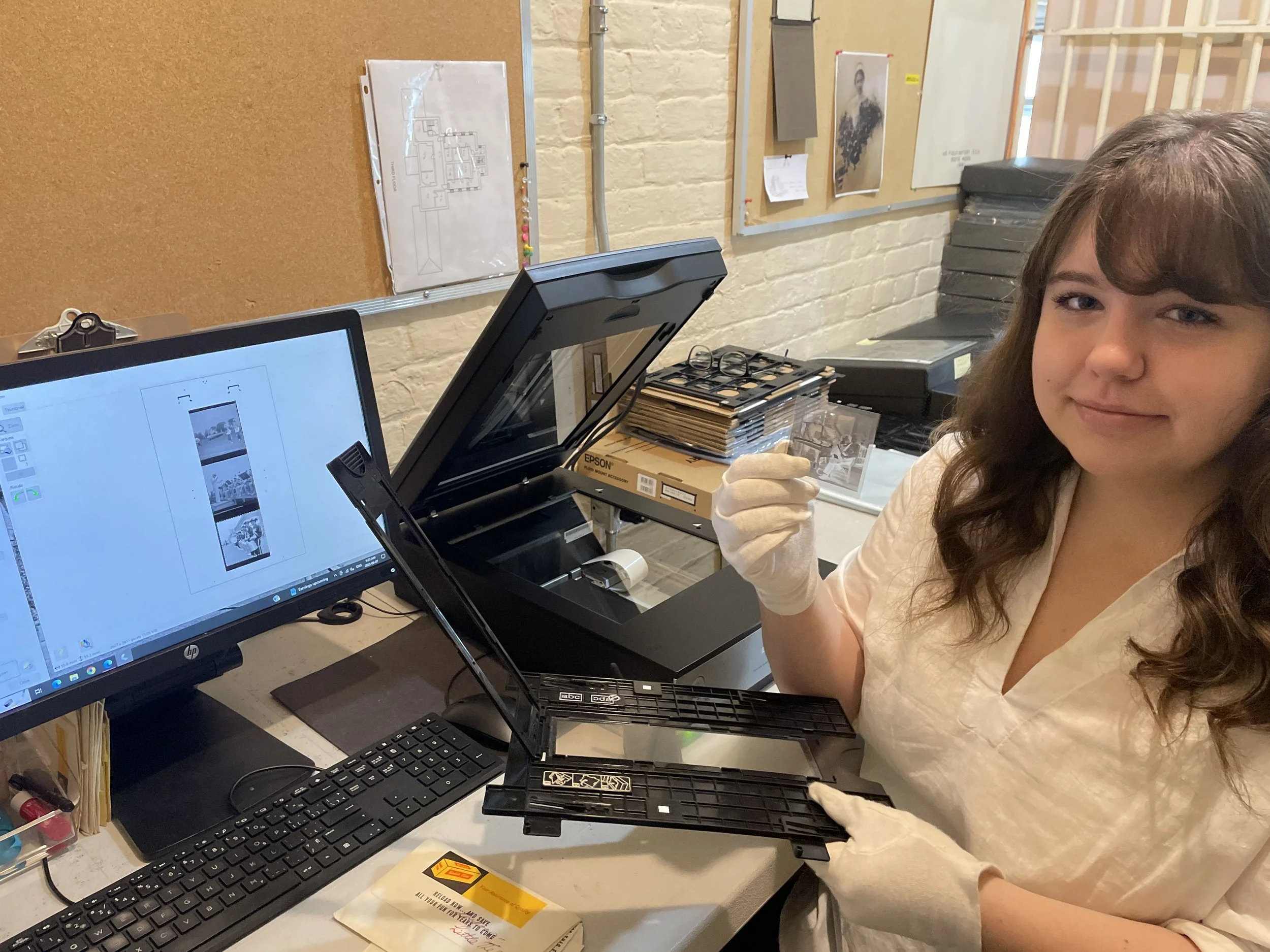 Woman in a white shirt and gloves holding a film negative in a workspace with a scanner and computer showing scanned images on screen.