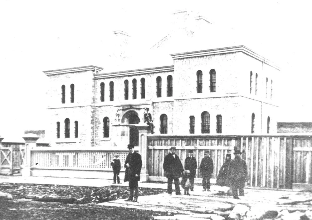 Black and white photo of a large 19th-century building with barred windows and several men standing in front, some in suits and hats, behind a wooden fence.