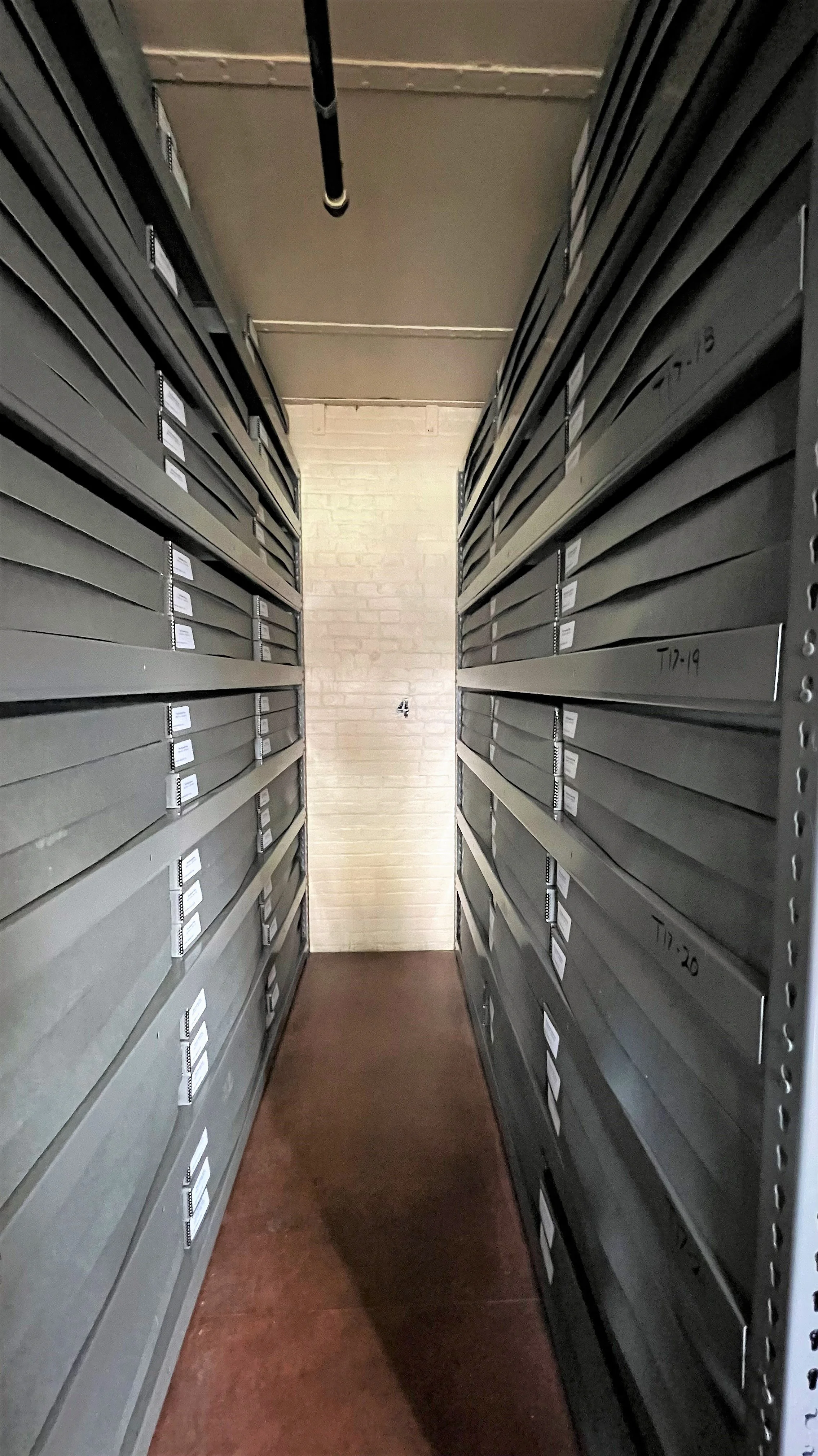 Narrow corridor with storage lockers on both sides, labeled with numbers and codes, in a dimly lit room.