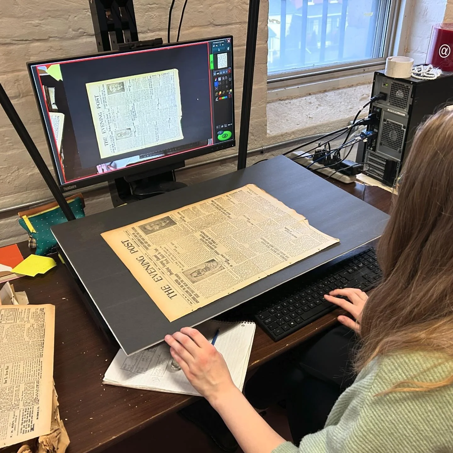Person digitizing a historical newspaper on a computer, with "The Evening Post" visible as the title on the paper. Image shows a computer setup with scanning equipment and a screen displaying the newspaper page.