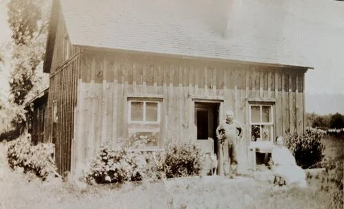Vintage photo of a rustic wooden house with two people standing outside in front of the building. The house has a steep roof and two windows on the front. A small garden with shrubs is visible in front of the house. The setting appears to be rural.