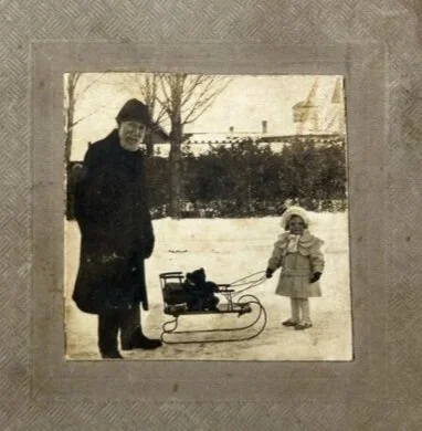 Vintage black and white photograph of an older man and a child in winter clothing pulling a sled in a snowy landscape, framed on textured paper with a metal hanger on top.
