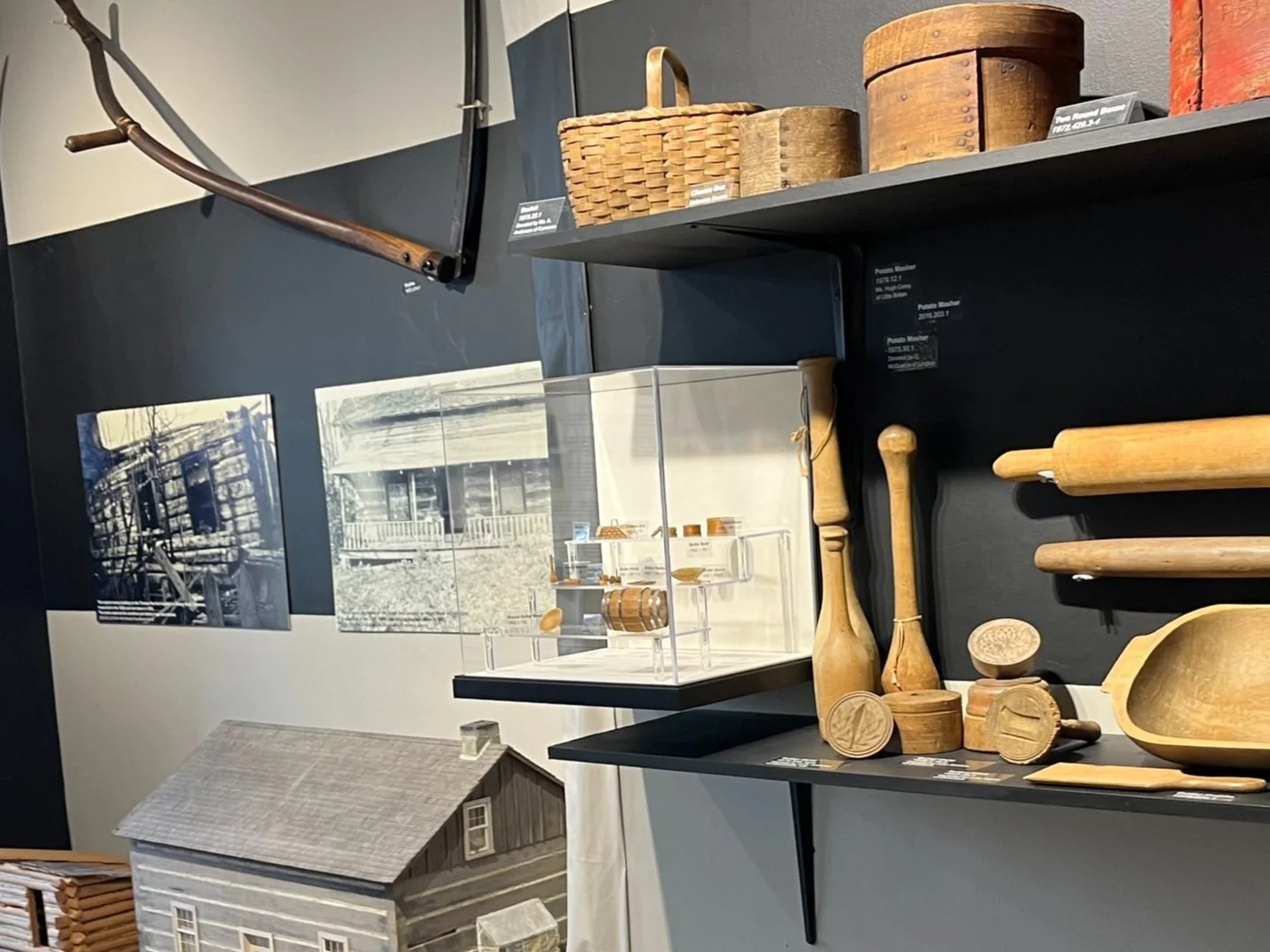 Museum display with antique kitchenware and wooden tools, including rolling pins, pestles, and a dough bowl. Baskets and wooden boxes are on the shelves. Historical photos and a model of a log cabin are mounted on the wall.
