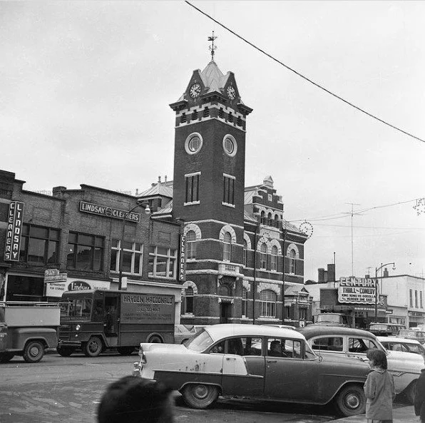 Vintage black and white photograph of a town square featuring a clock tower and parked cars, likely from the mid-20th century. Surrounding buildings include shops displaying signs for cleaners, hardware, and a theater.