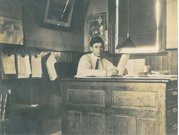 Charles Heels as a young man sitting at a desk in the GTR Railway Station. Image is B&W.