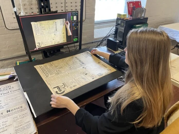 Woman scanning a newspaper page using a large flatbed scanner with a computer monitor displaying the scanned image in a workspace.