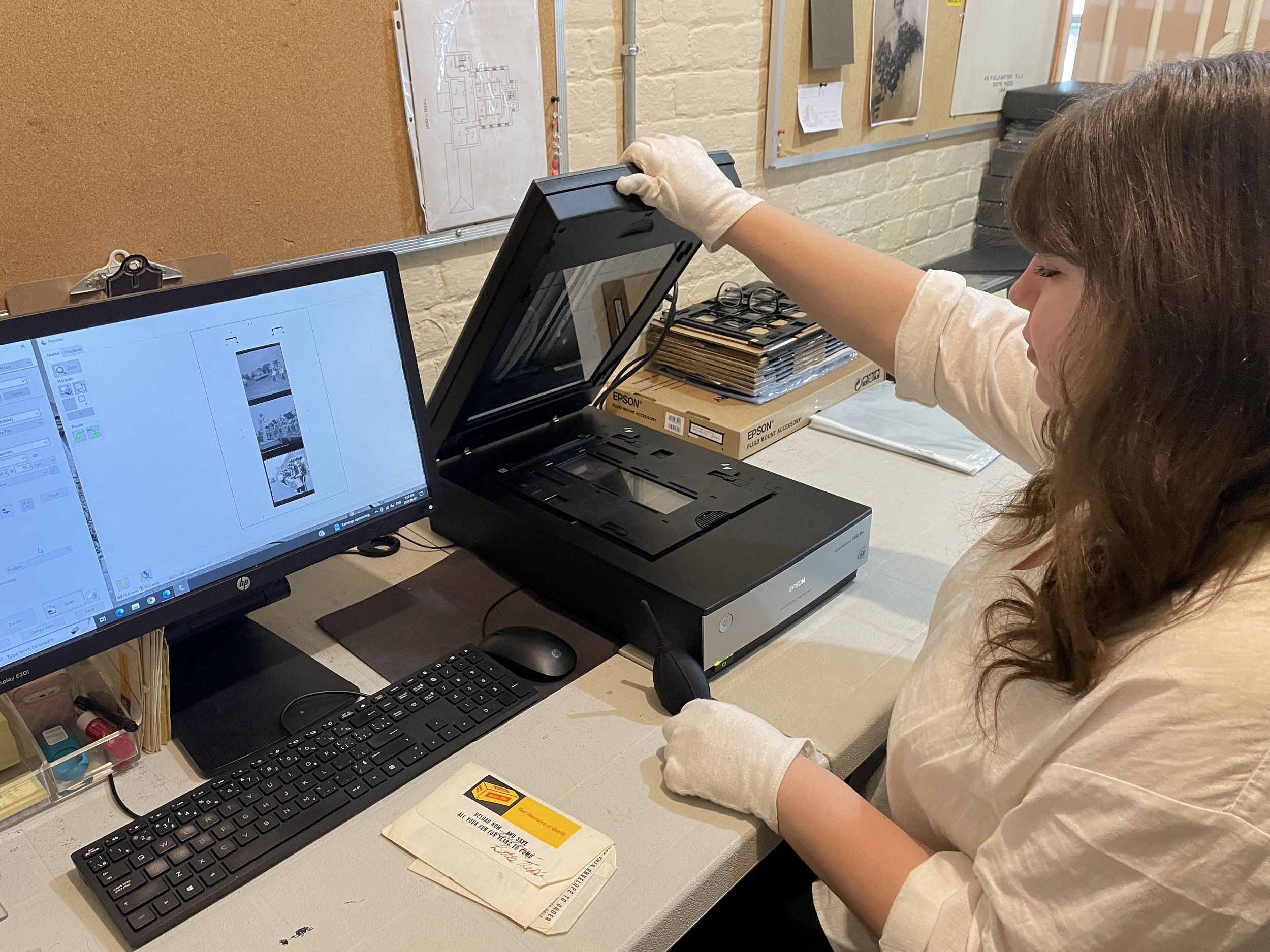 Woman scanning photo negatives with a flatbed scanner near a computer monitor displaying scanned images, wearing white gloves.