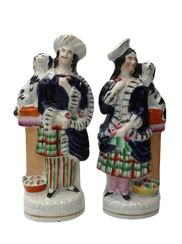 Antique Staffordshire-style ceramic figurines depicting a man and a woman in colorful 18th-century attire with dogs at their sides, standing on decorative bases.