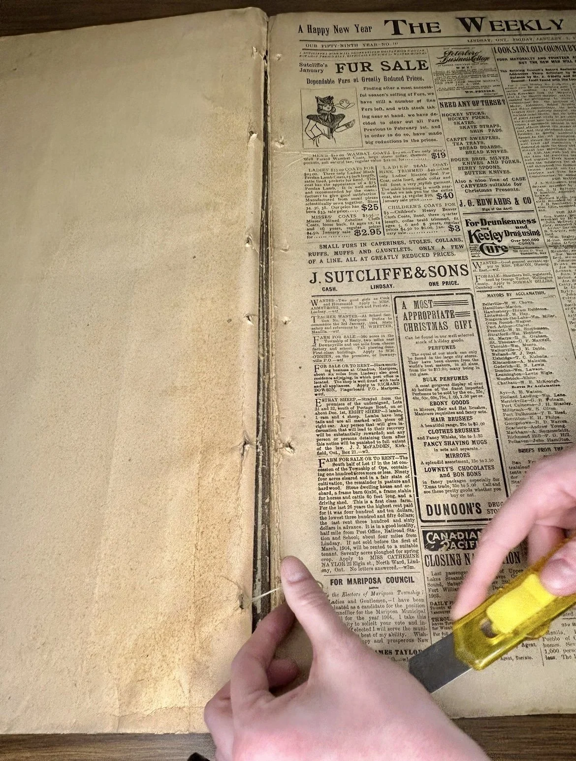 Hands using a box cutter to cut along the spine of an old newspaper titled "The Weekly," featuring vintage advertisements and text.