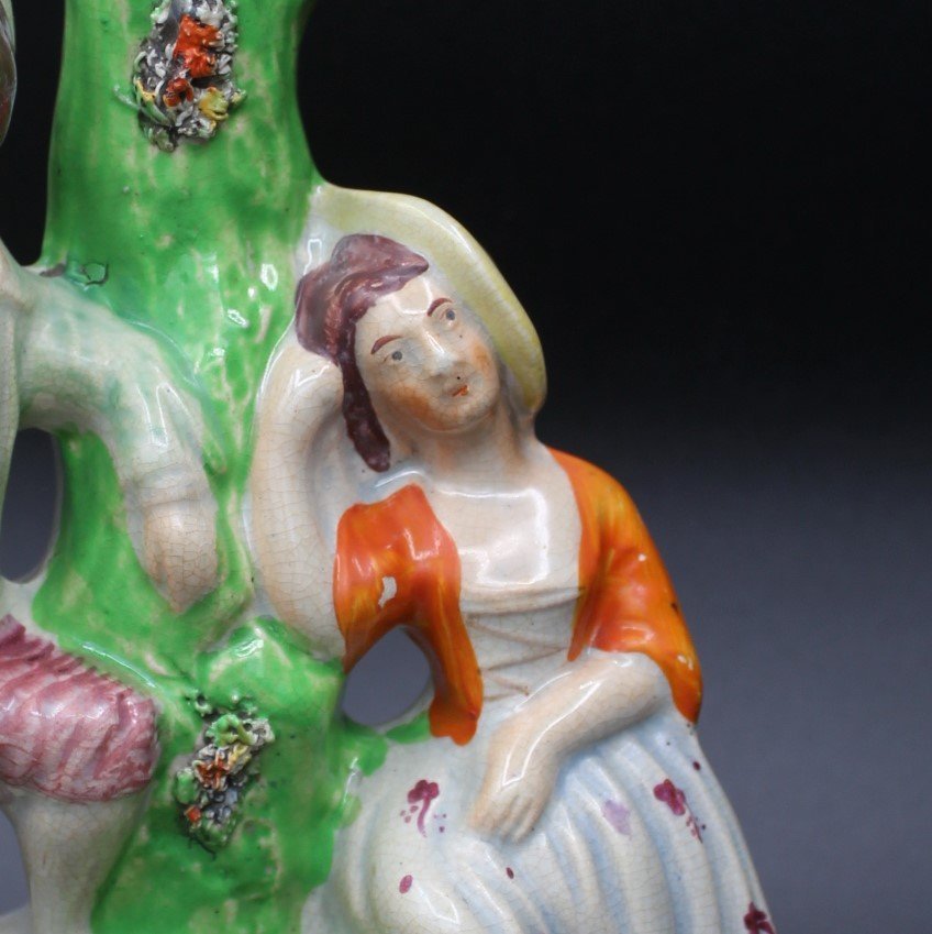 Close-up of a ceramic figurine depicting a woman in a colorful dress with an orange shawl, leaning against a green tree. The figurine shows intricate detailing in the face and attire.
