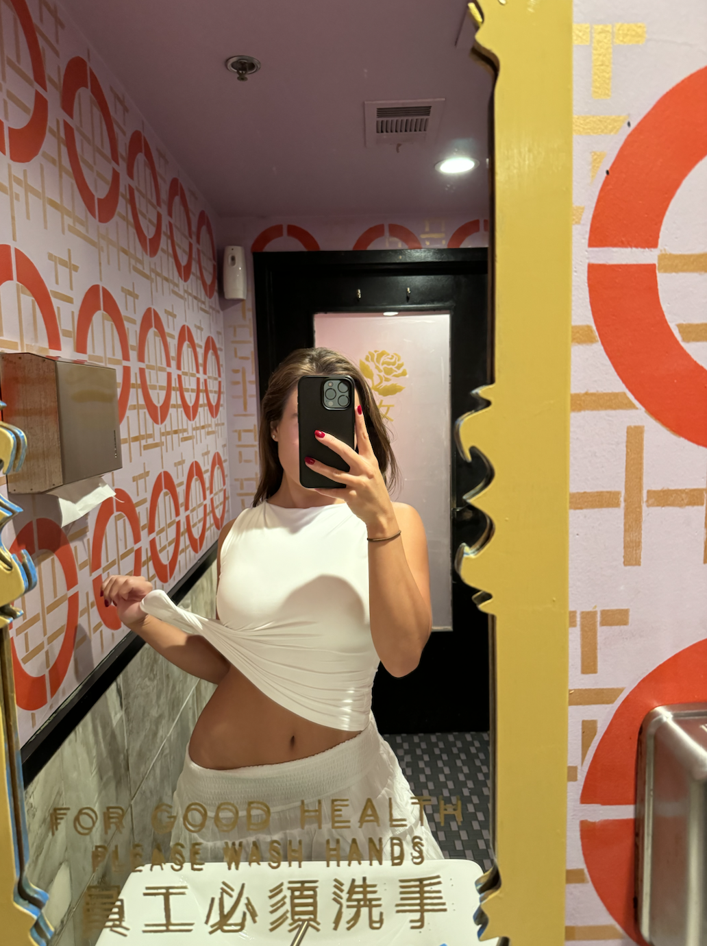 Arabella Blair, Independent Escort in London, taking a mirror selfie in a colorful bathroom. She is wearing a white sleeveless top and beige shorts, holding her top up with one hand. 