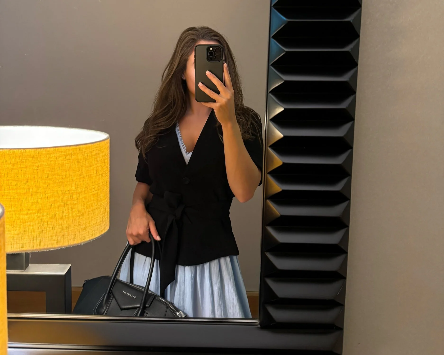 Arabella Blair, Independent Escort in London, taking a mirror selfie in a hotel dressing area, holding a black handbag, wearing a black top and a skirt, with a yellow lampshade and a decorative black panel in the background.