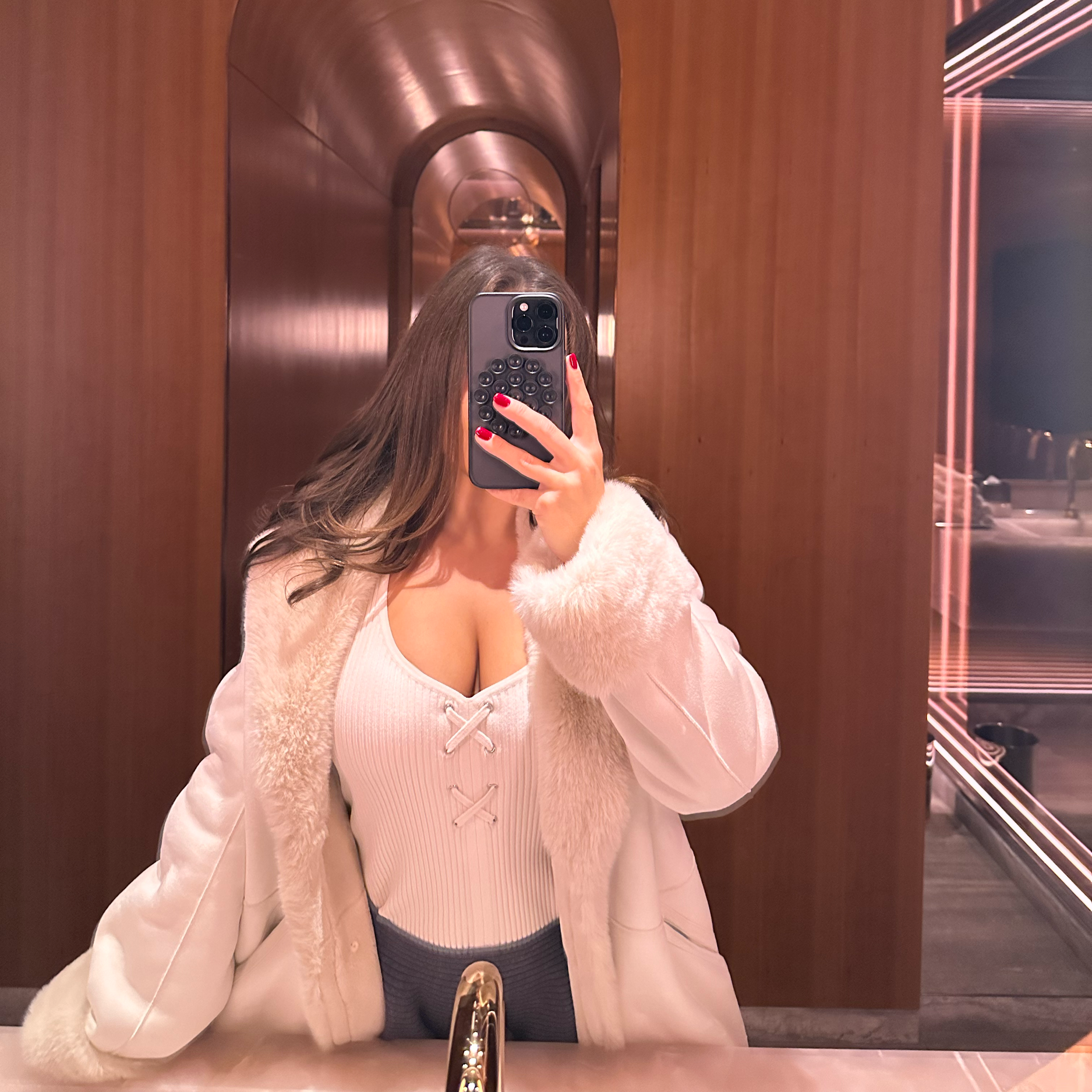 Arabella Blair, Independent Escort in London, taking a mirror selfie, wearing a cream-colored jacket with fur cuffs, a beige top with lace-up detail, and dark skirt.
