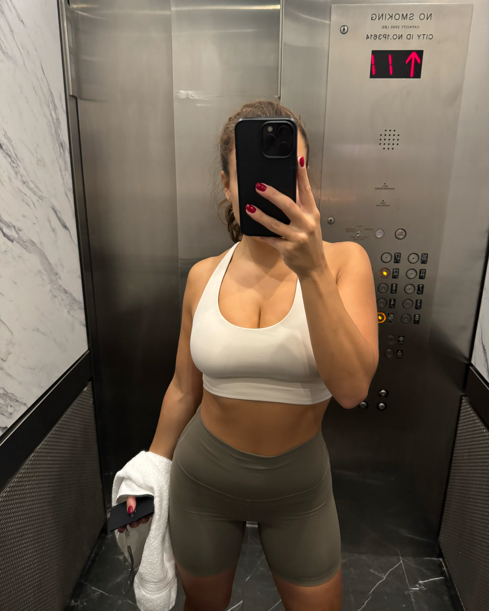Arabella Blair, Independent Escort, taking a mirror selfie in an elevator, wearing a white sports bra and olive green shorts. She has long brown hair pulled back, red nail polish. The elevator interior is metallic with control buttons visible.